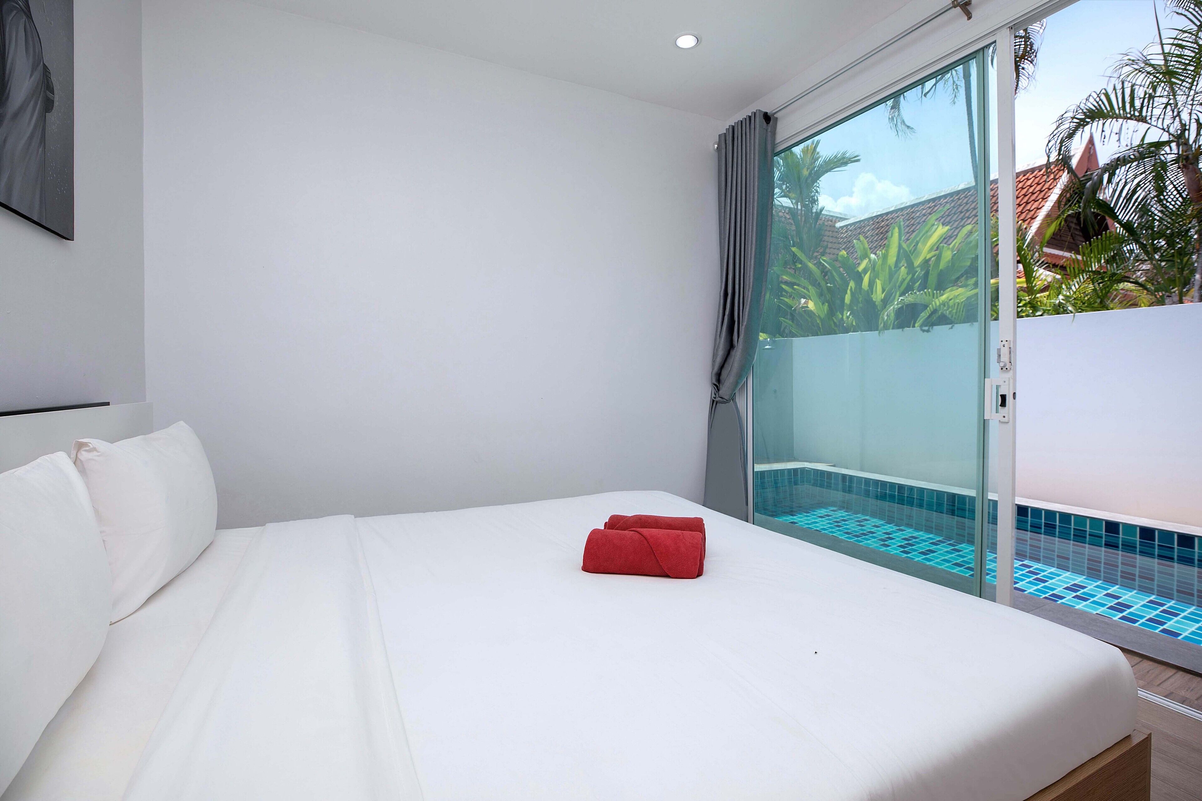 A hotel room with a bed, a red towel, a glass door leading to a pool area, and a view of greenery outside.