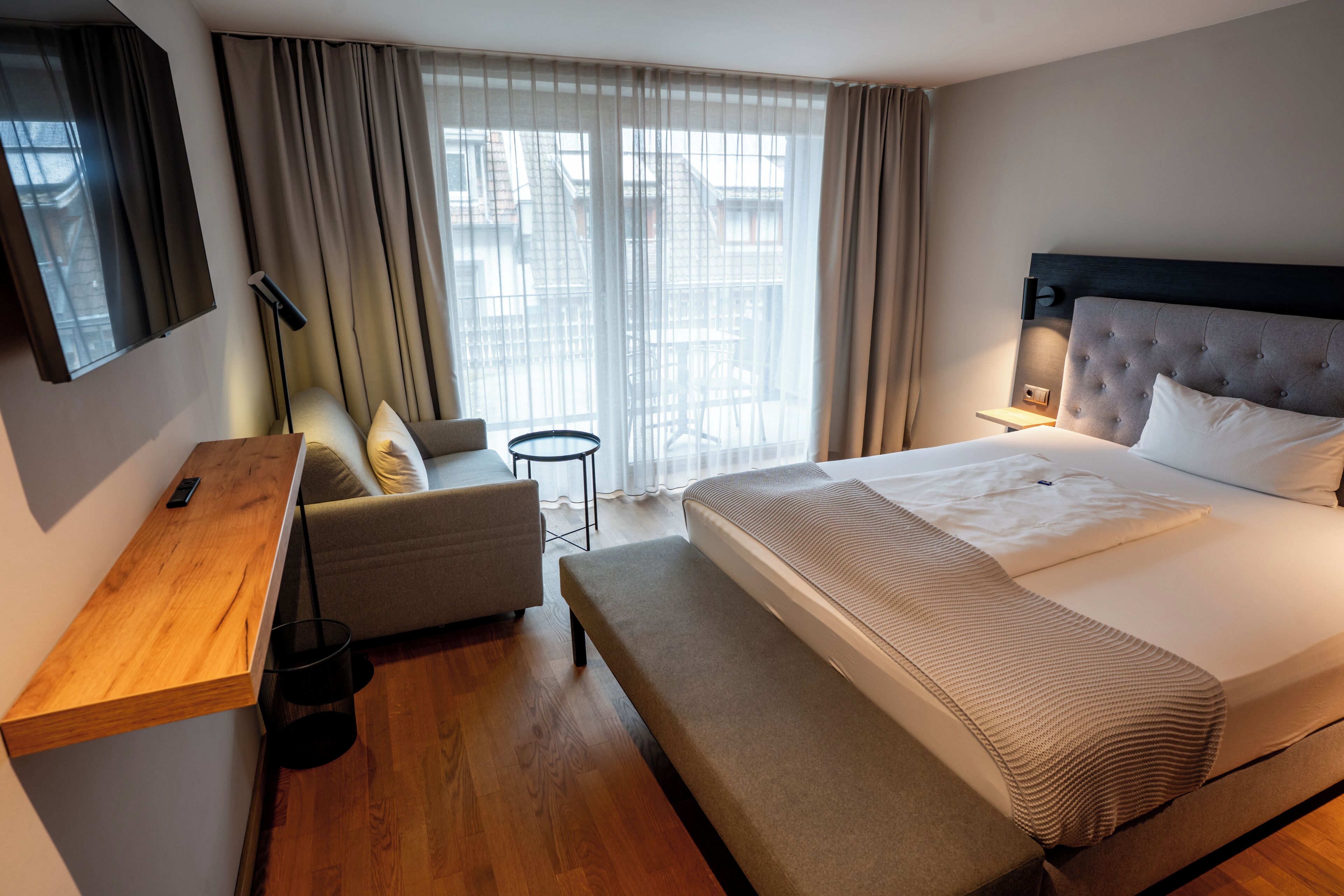 A modern hotel room with a large bed, a wooden desk, a chair, a small table, and a TV mounted on the wall.