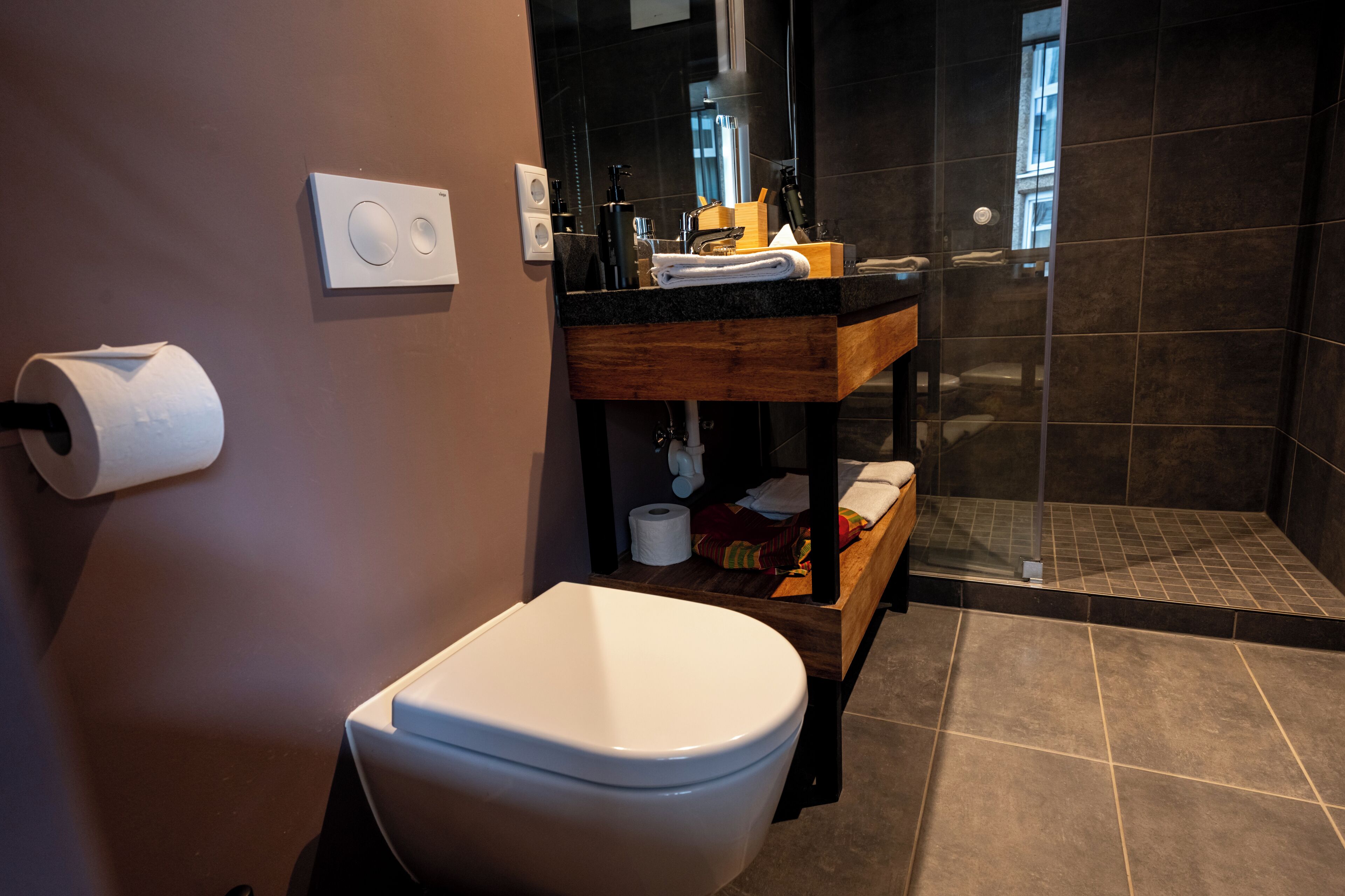 Ein modernes Badezimmer mit Toilette in Weiß, einem Holzwaschtisch, einer Duschkabine aus Glas und einem dunklen Fliesenboden.
