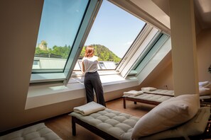 Una habitación con tragaluz, cama, banco y vistas a una colina con árboles.
