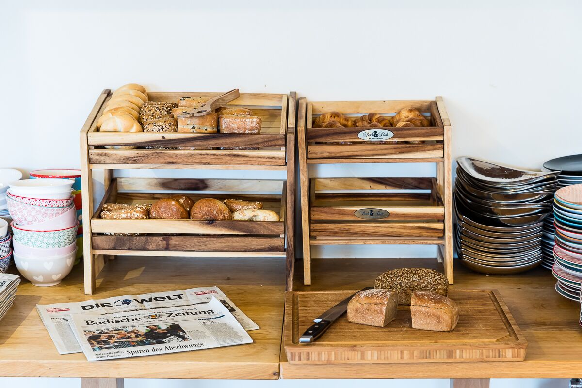Wooden bread racks with various breads and pastries, a newspaper, and stacked plates on a table.