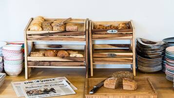Wooden bread racks with various breads and pastries, a newspaper, and stacked plates on a table.
