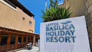 La entrada al Leptos Basilica Holiday Resort, con un letrero, zona de estar al aire libre y fachada de edificio.