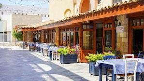 Un área de comedor al aire libre con mesas y sillas, piso de baldosas y un edificio con techo de tejas y puertas de madera.