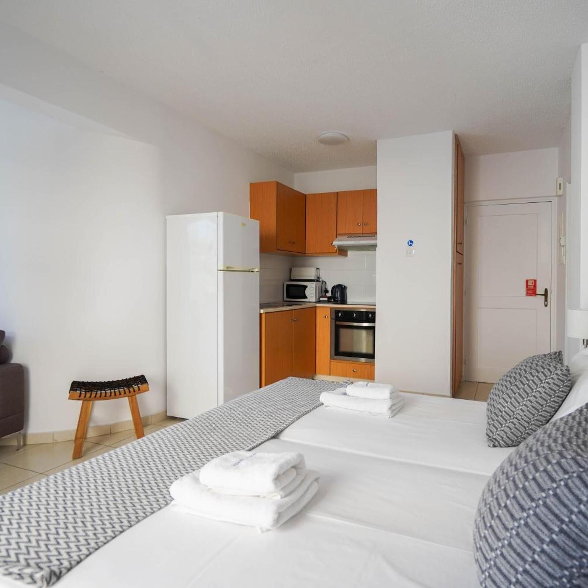 Una habitación de hotel compacta con kitchenette, una cama y toallas dobladas con cuidado, además de un taburete pequeño.