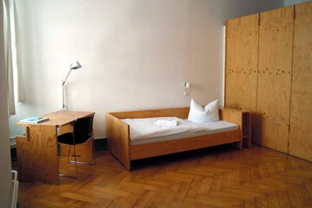 A small, single-bed room with a wooden desk and chair, a bedside lamp, and a wardrobe.