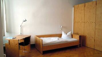 Una habitación pequeña con una cama individual, un escritorio y una silla de madera, una lámpara de noche y un armario.