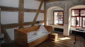 Ein hölzernes Bett mit weißer Matratze und Kissen, ein kleiner Nachttisch aus Holz sowie ein Fenster mit Blick ins Freie.