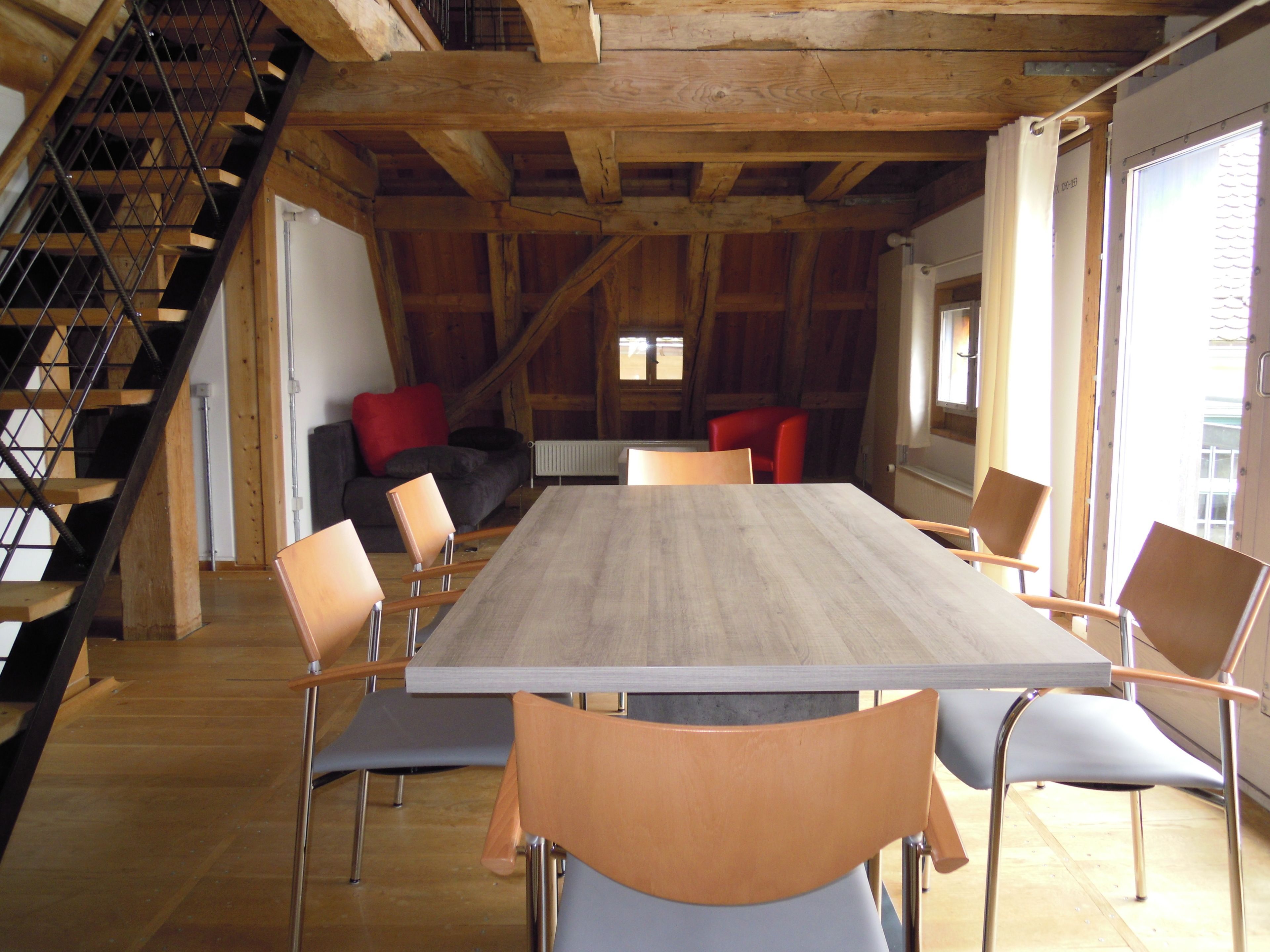 A spacious room with a large wooden table, several chairs, a staircase, and a cozy seating area with cushions.