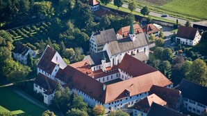 Ein Luftbild eines historischen Gebäudekomplexes mit mehreren Gebäuden, darunter eine markante Kirche mit Turm, umgeben von üppiger Vegetation und offenen Feldern.