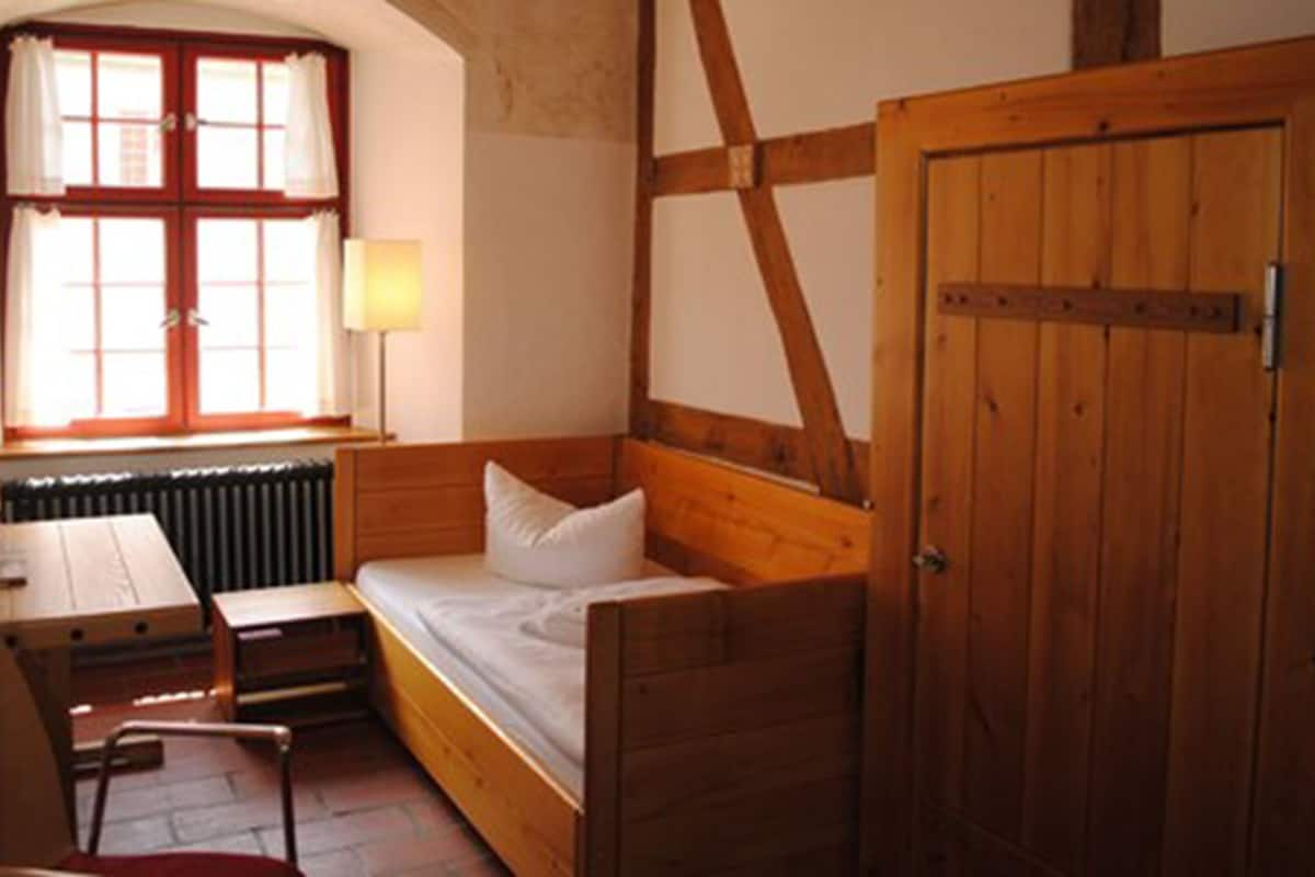 A small, cozy room with a wooden bed, a desk, and a chair. There is a window with curtains and a radiator below it.