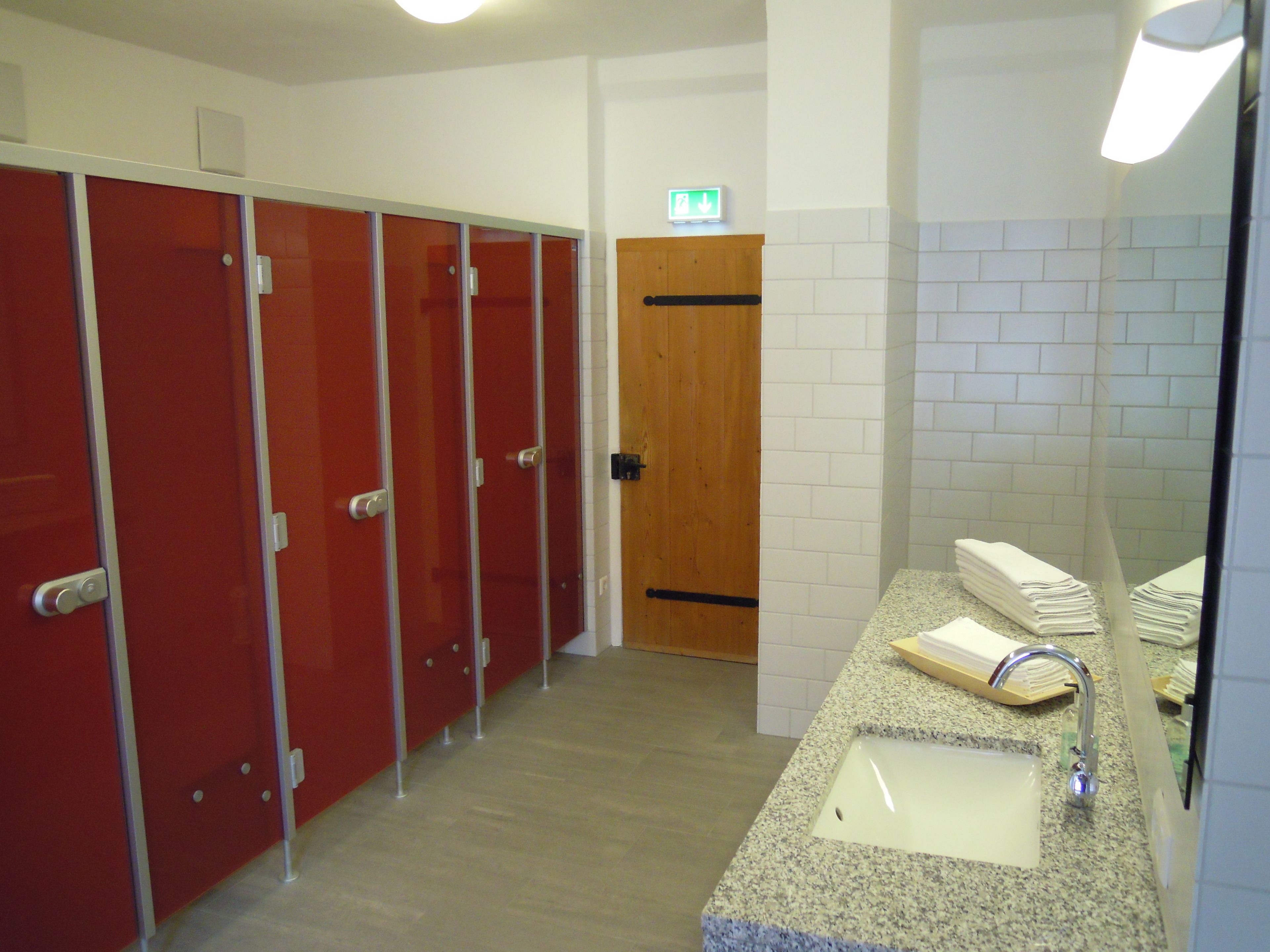 Un baño público con cabinas rojas, una puerta de madera, un lavabo con espejo y un perchero para toallas.