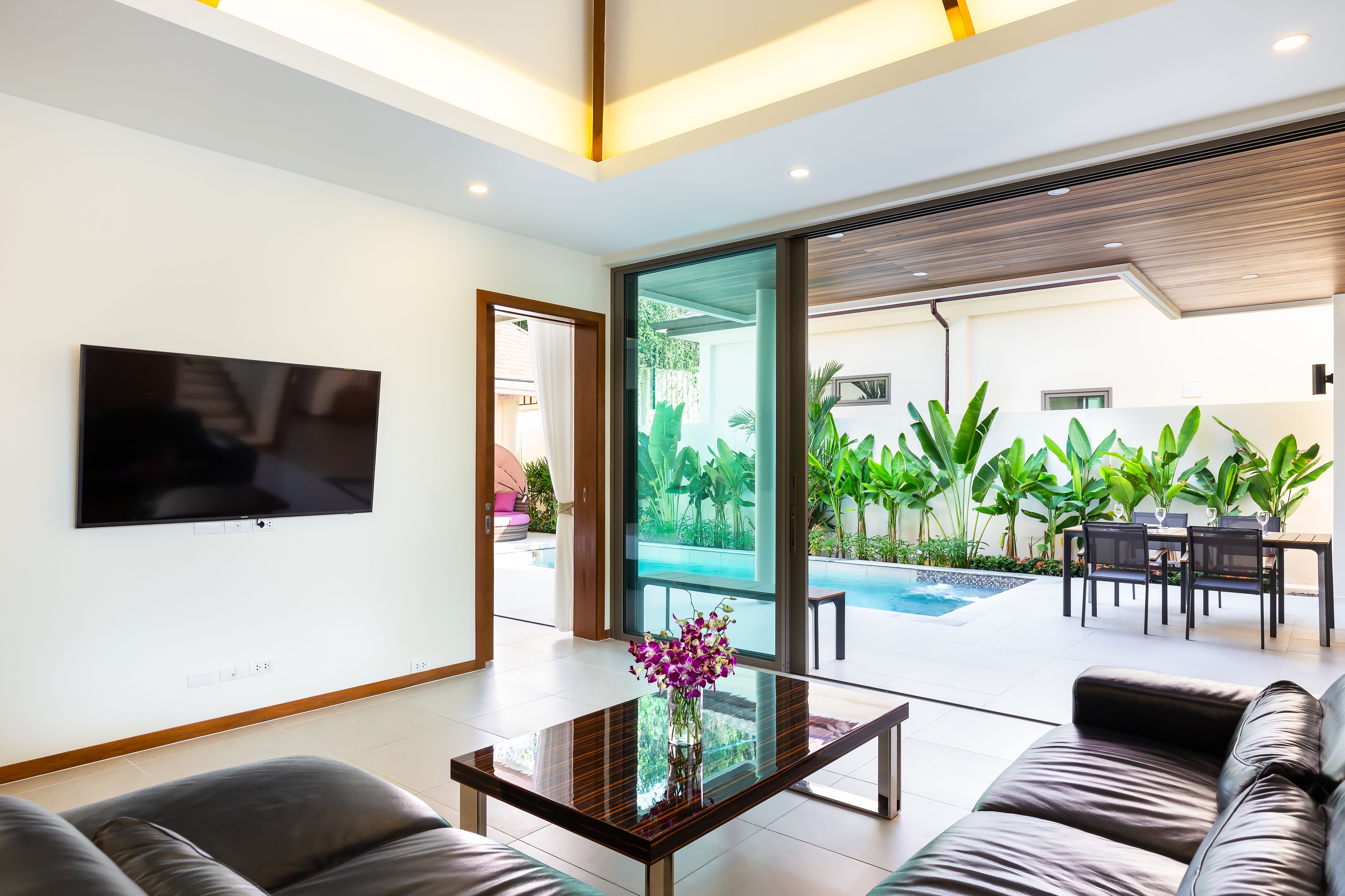 A modern living room with a flat-screen TV, a glass coffee table with flowers, a leather sofa, and a view of a pool and outdoor dining area.