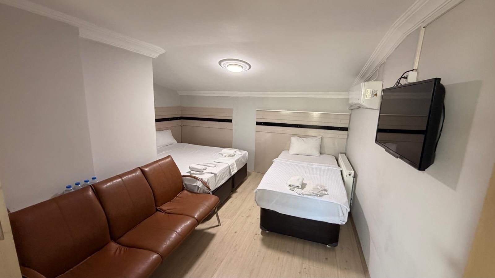 Quarto de hotel pequeno com sofá de couro marrom, duas camas de solteiro, TV de tela plana e piso de madeira.