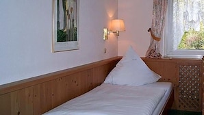 Una cama individual con cabecera de madera, una lámpara de pared, un cuadro enmarco y una ventana con cortinas.