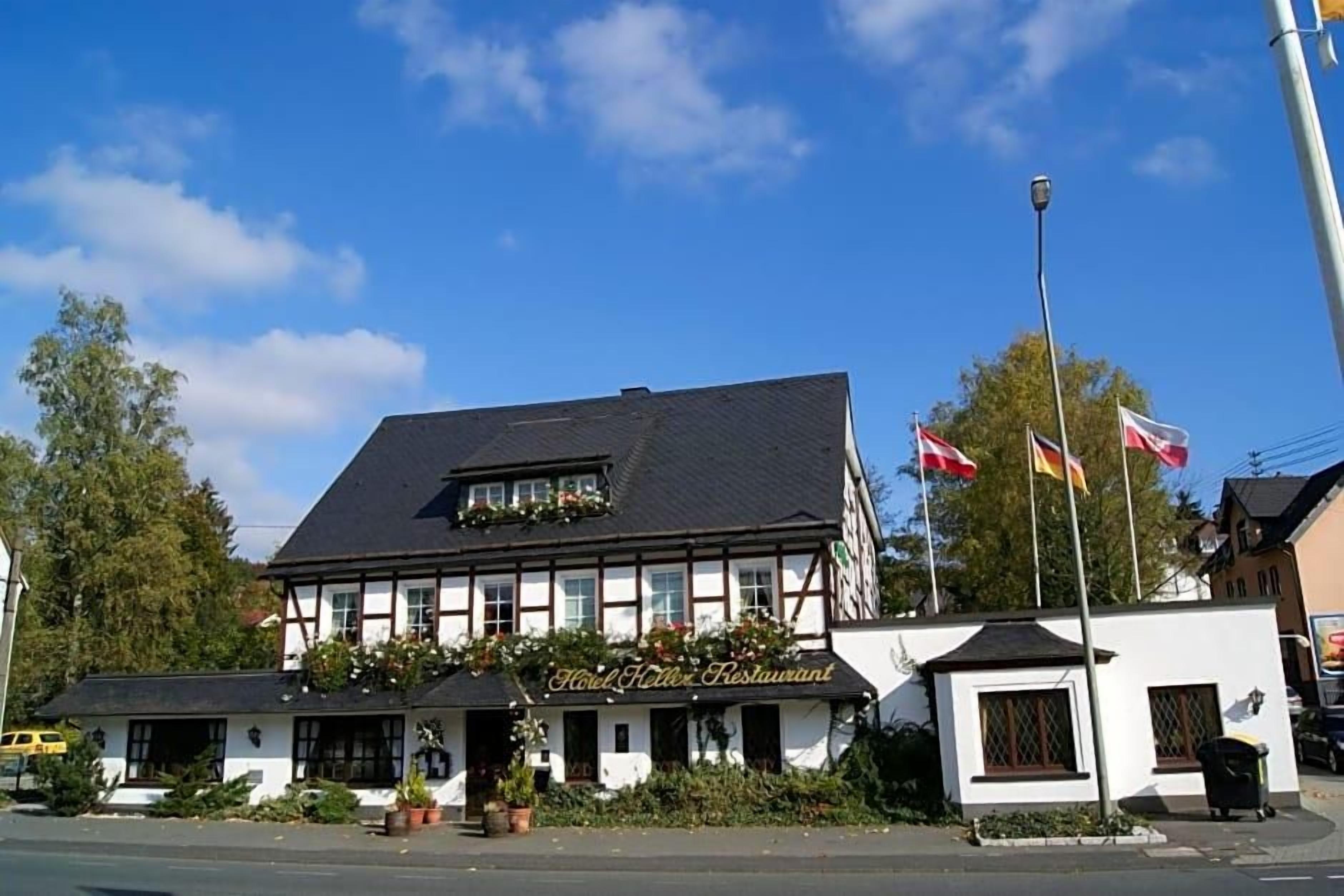 Ein traditionelles Gebäude mit einem Schild, auf dem 'Hotel Keller Restaurant' steht, ein kleiner Anbau und Flaggen an einem Mast.