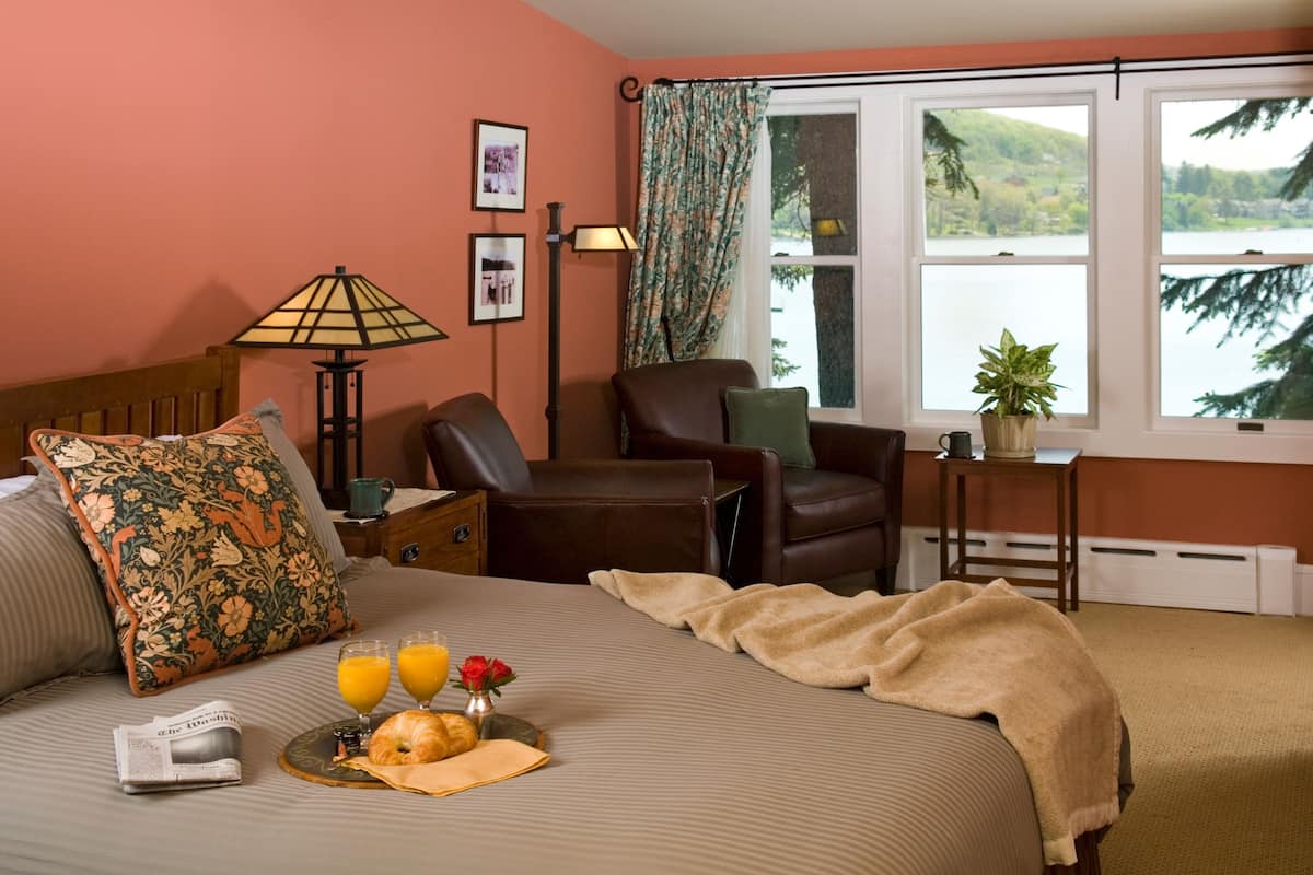 A hotel room with a bed, a chair, a table with a plant, and a window with a view of a lake.