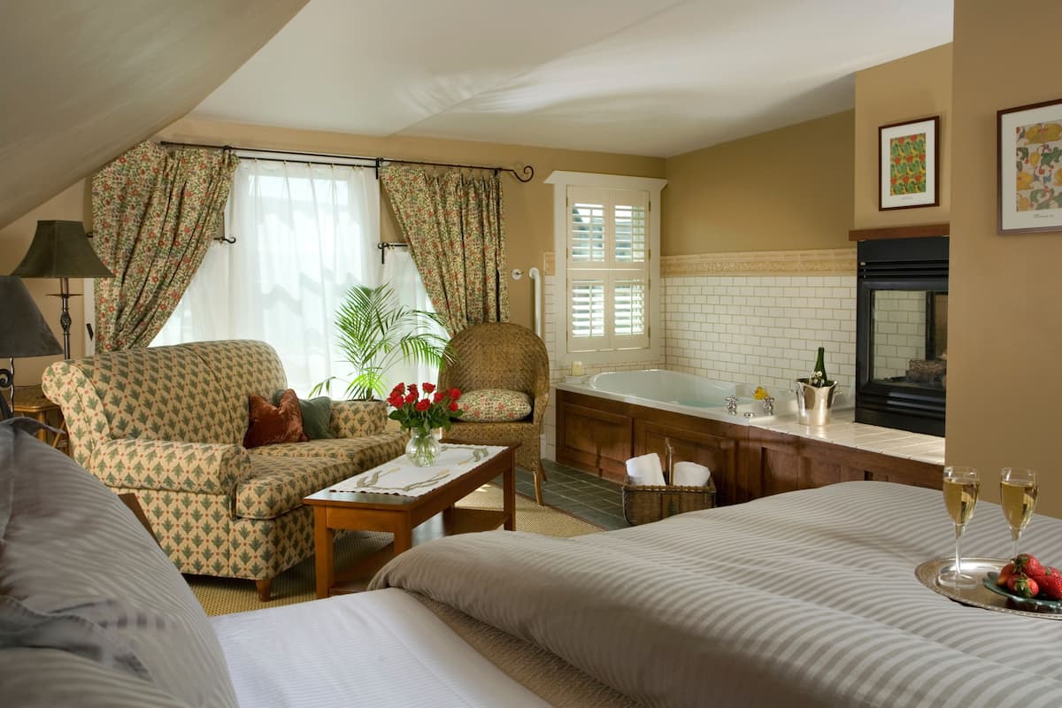 A hotel room with a sofa, armchair, a small table with a vase of flowers, a bathtub, and a tray with strawberries and champagne glasses.