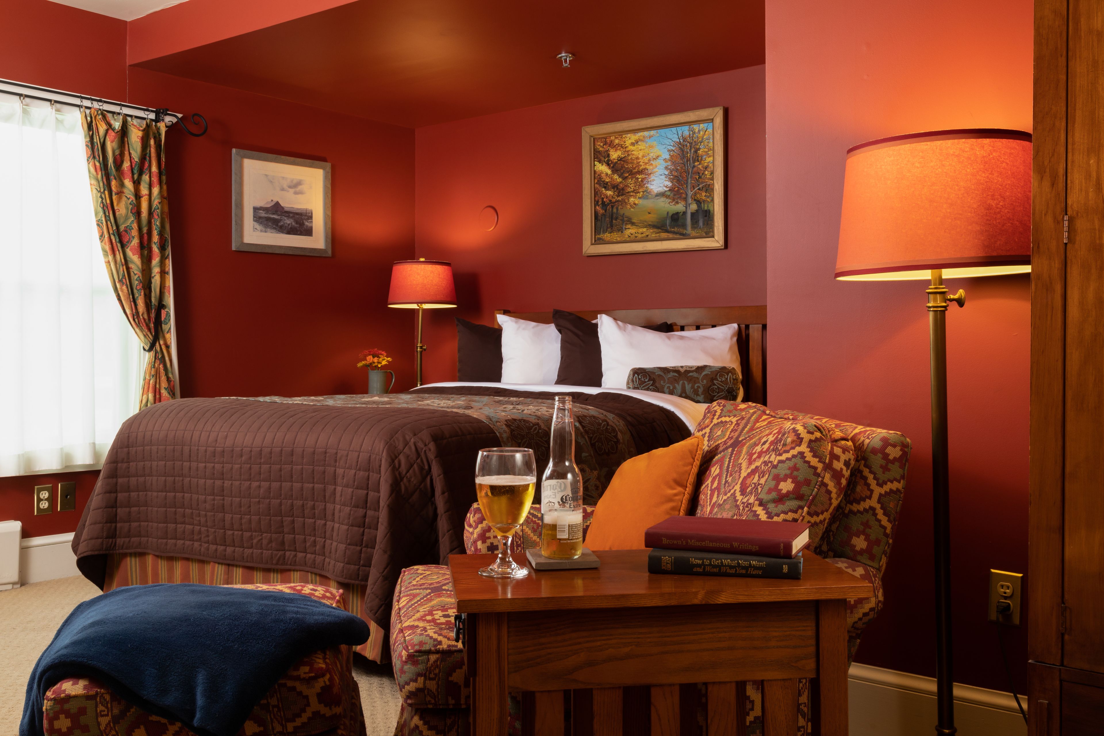 A hotel room with a bed, a bedside table with a glass of beer and a book, a lamp, and a painting on the wall.