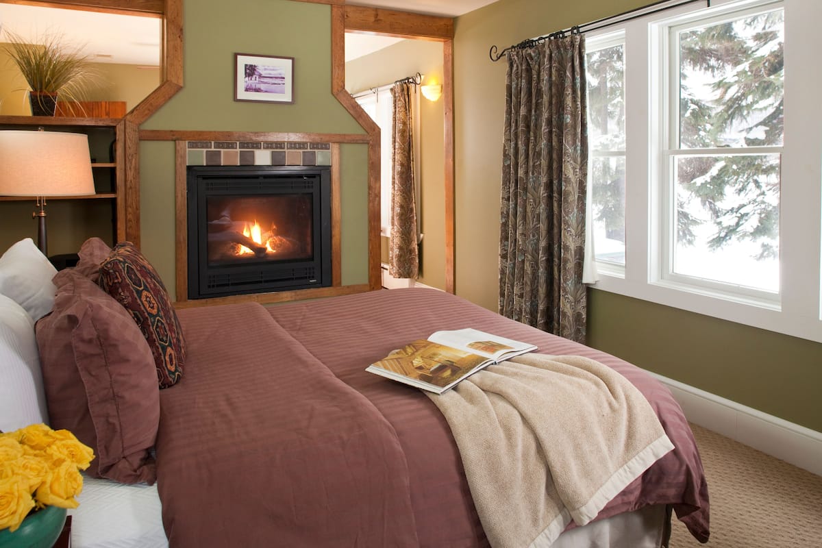A bedroom with a fireplace, a bed with a red blanket, a window with curtains, and a view of trees outside.