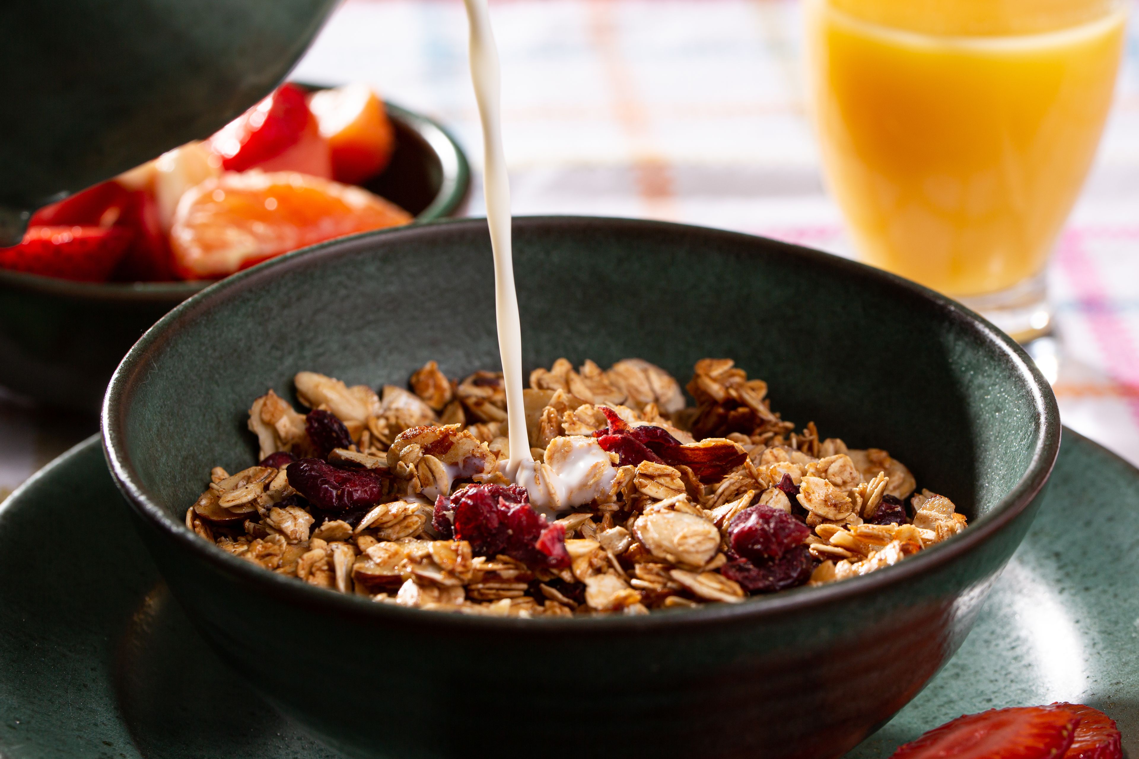 Un bol de granola dans lequel on verse du lait, posé sur une table. On aperçoit en arrière-plan un verre de jus d'orange.