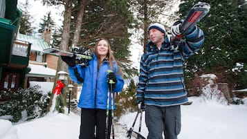Two people with skis standing in a snowy outdoor area with trees and a building in the background.