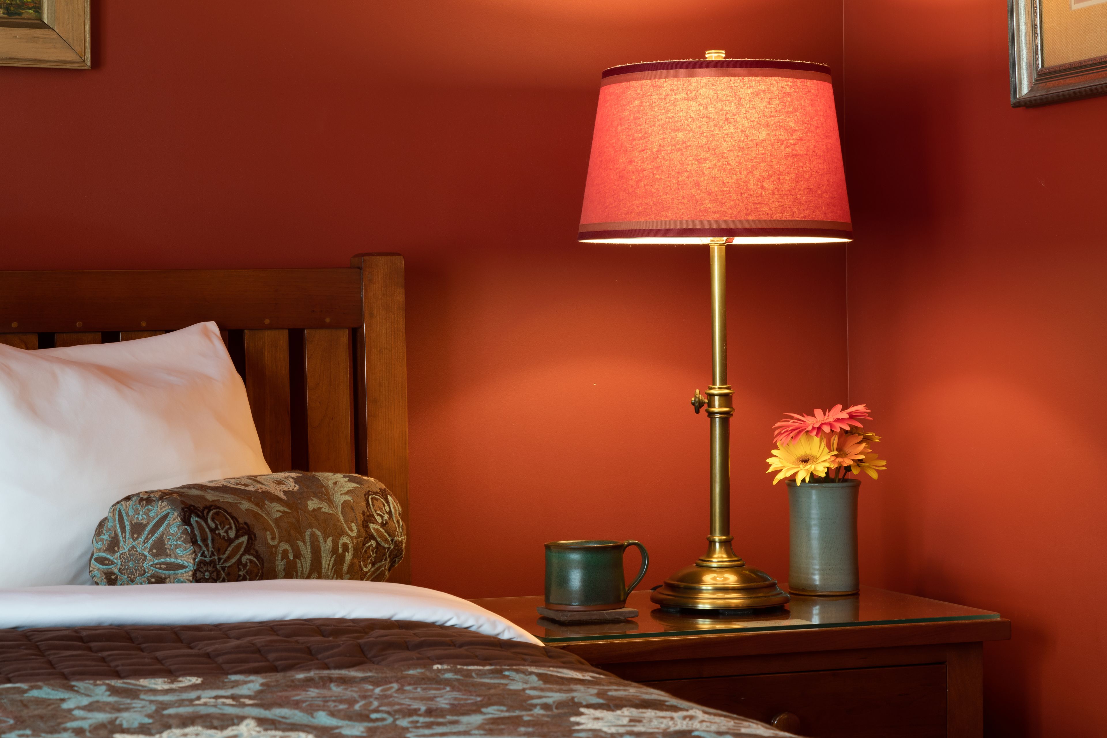 A neatly made bed with a patterned pillow, a bedside table with a lamp, a vase with flowers, and a mug.