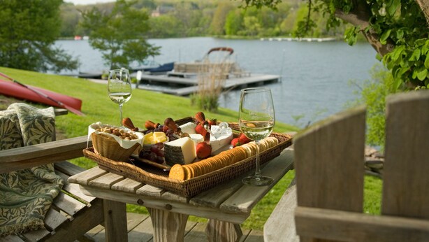 A picnic setup with a wicker basket containing cheese, grapes, and strawberries, and two glasses of white wine on a wooden table overlooking a lake. - Lake Pointe Inn (McHenry)