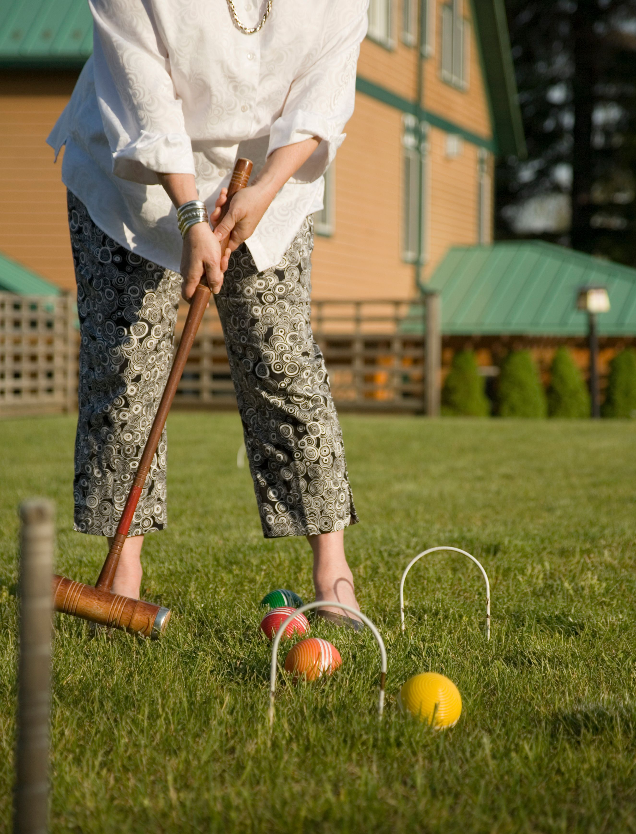 Henkilö pelaa krokettia ruohikkoisella kentällä, taustalla näkyy talo.