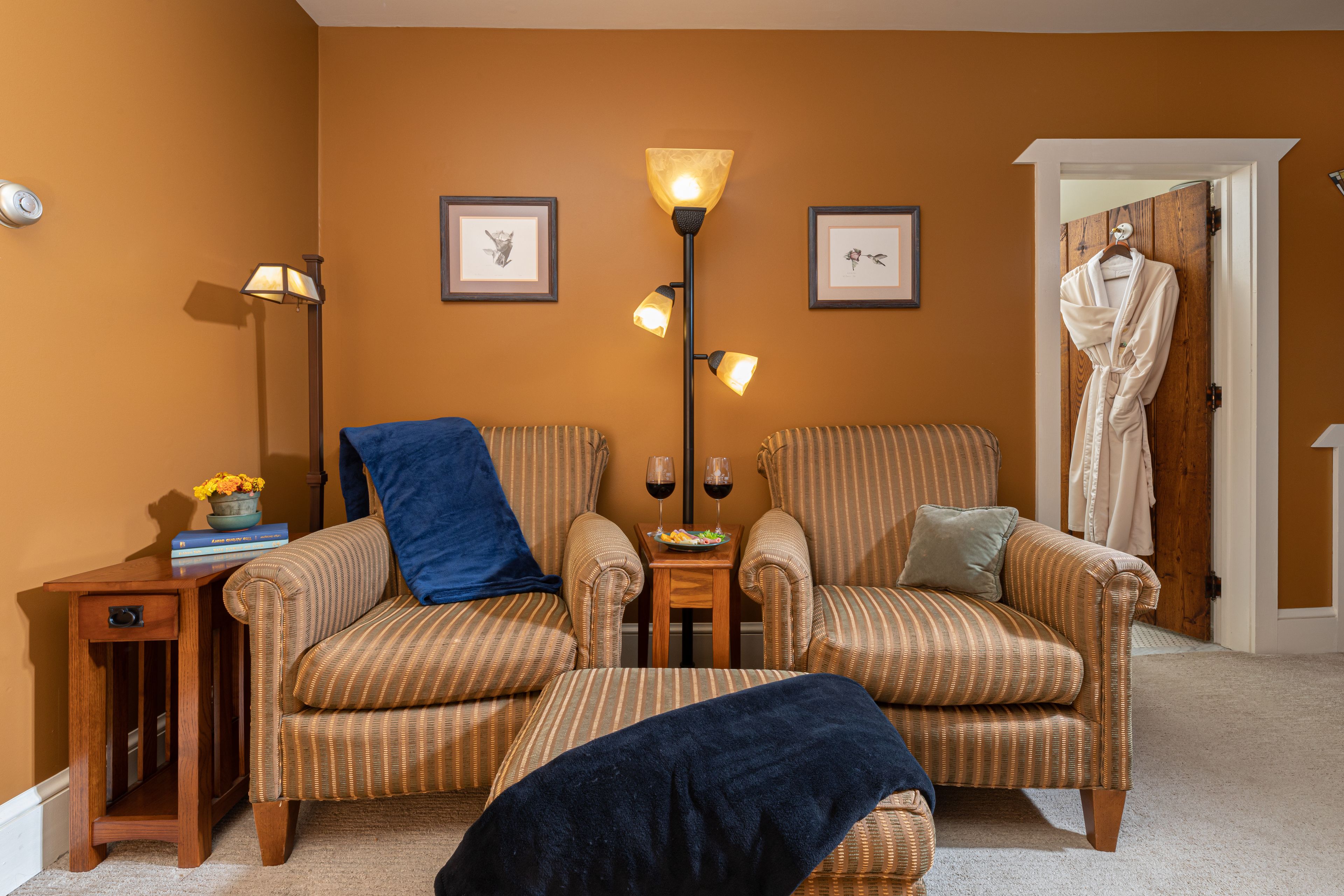 A cozy room with striped armchairs, a small side table with a lamp, a robe hanging on the door, and framed pictures on the wall.