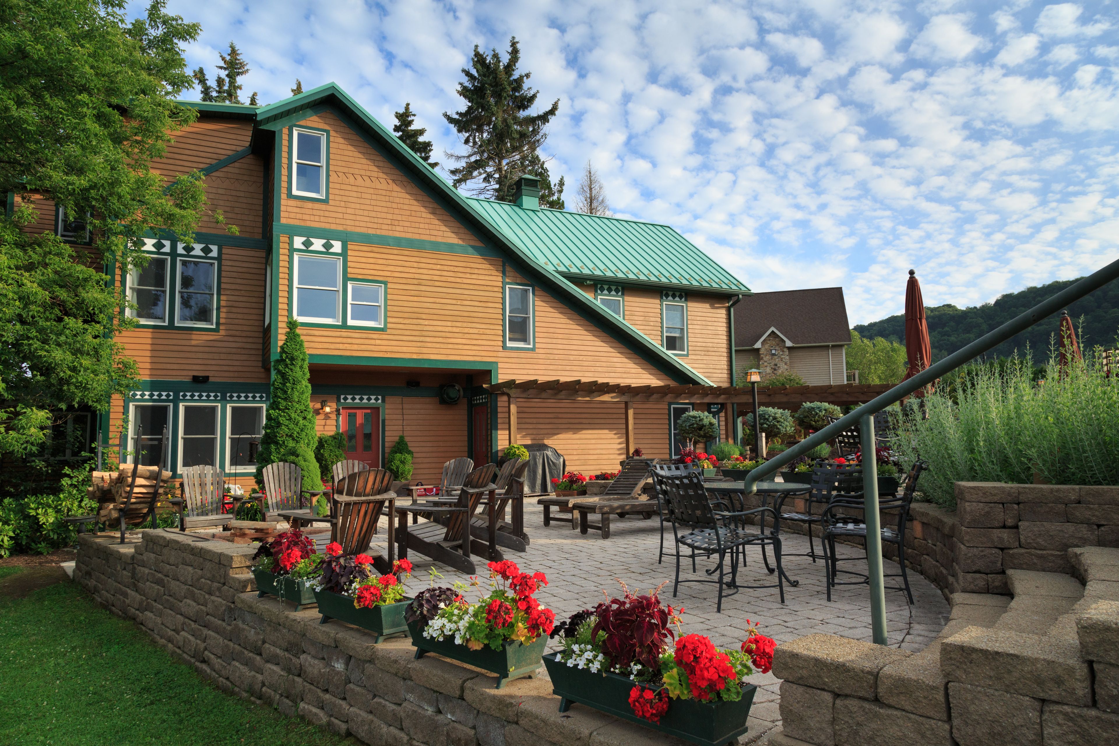 A two-story wooden building with a green roof, outdoor seating area with chairs and tables, and a stone retaining wall with flower planters.