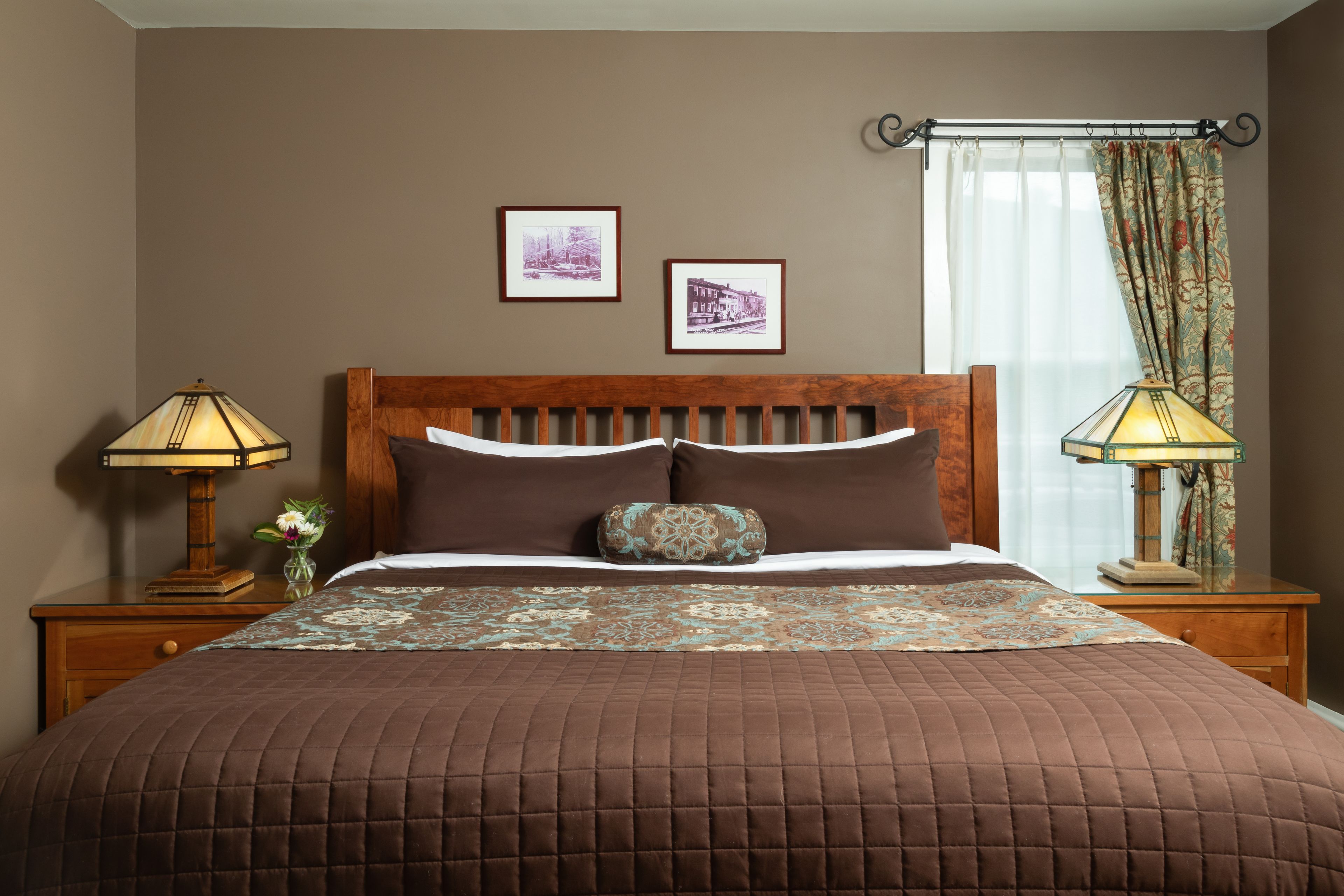 A neatly made bed with a brown quilt, two framed pictures on the wall, and a window with curtains.