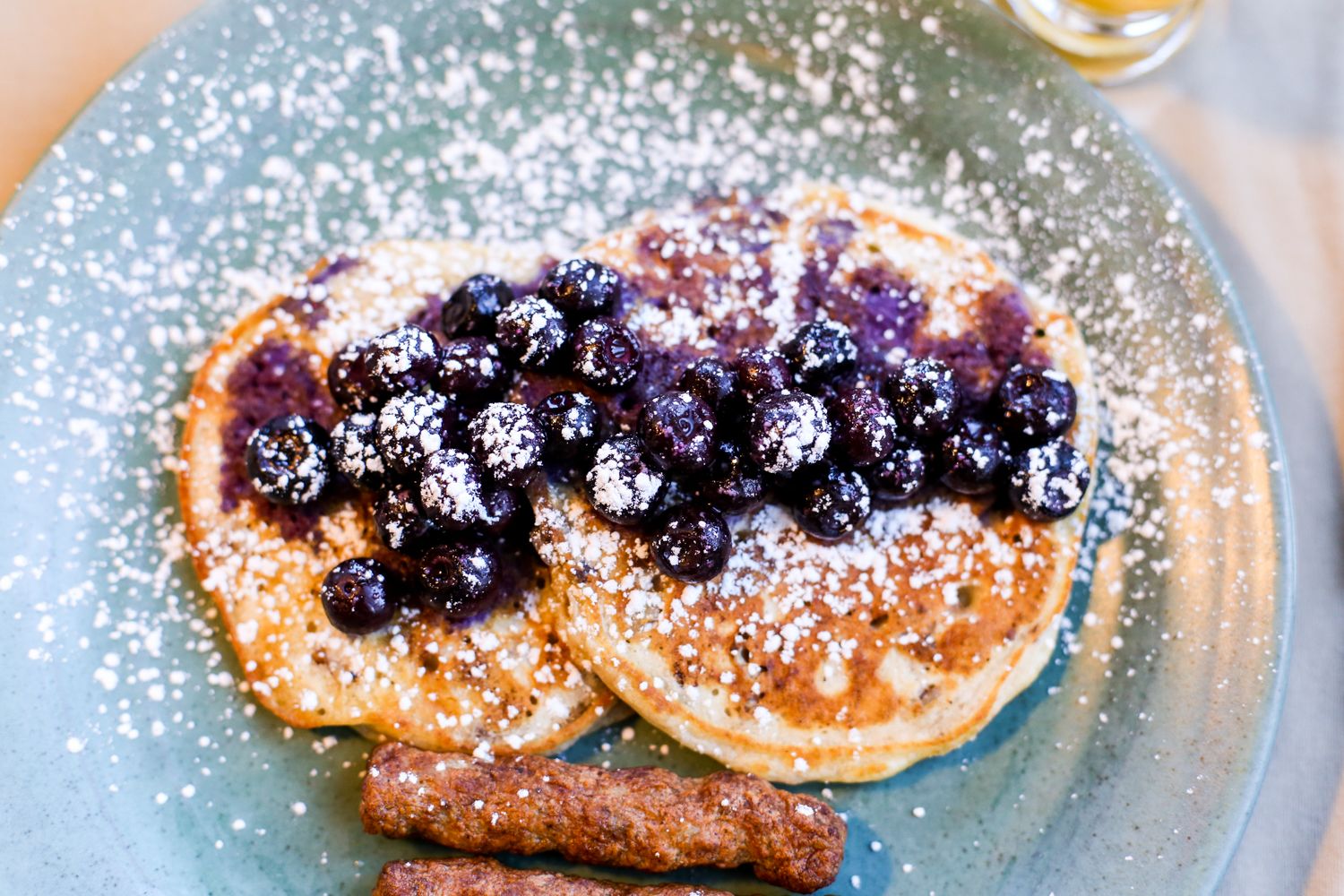Un plat amb crepes cobertes de blueberries i sucre glas, acompanyat d'una salsitxa.