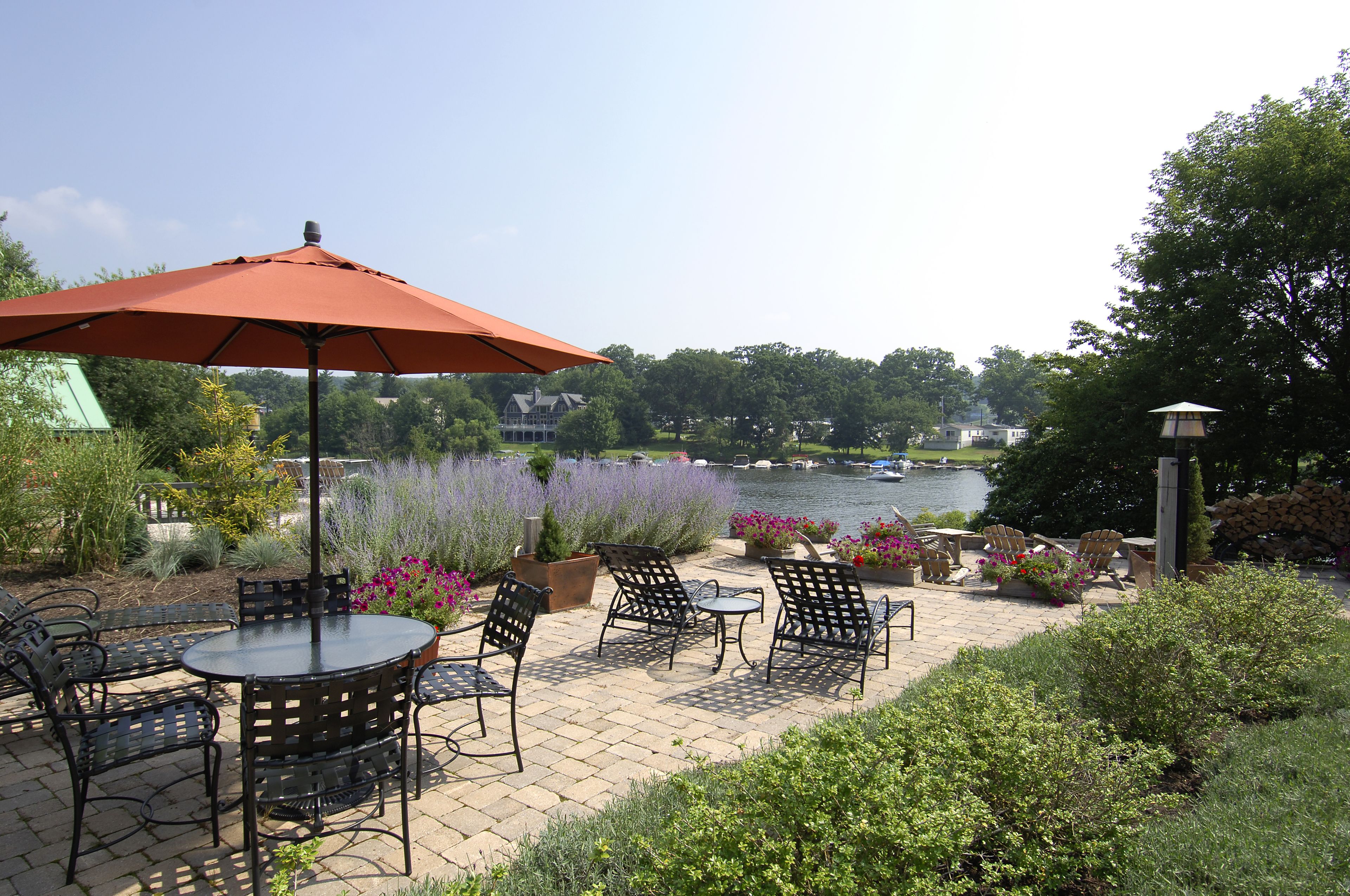 Une terrasse extérieure avec une table ronde et des chaises, un grand parasol et une vue sur un lac où naviguent des bateaux.