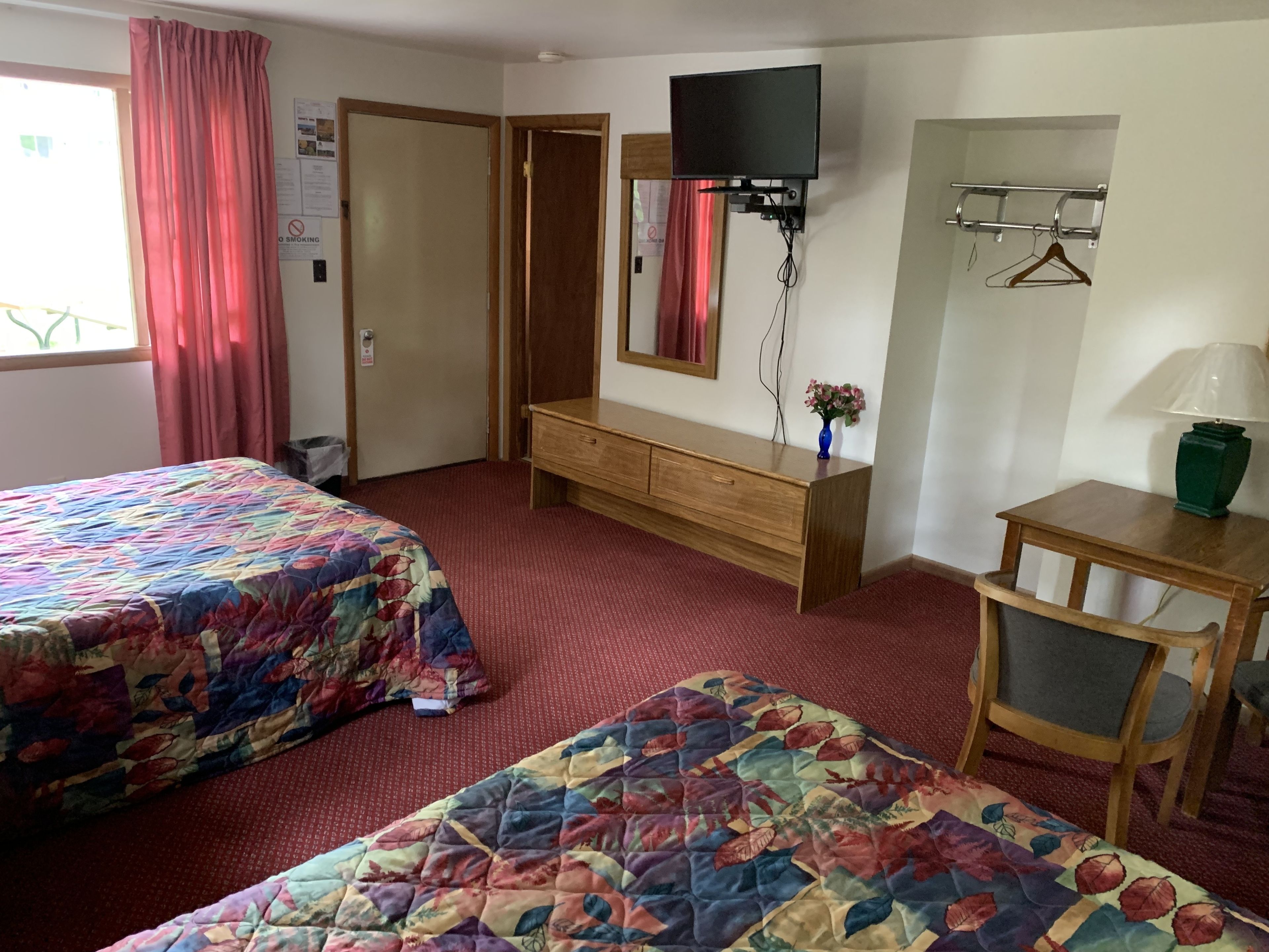 A hotel room with two beds, a wooden dresser, a television mounted on the wall, and a desk with a lamp.