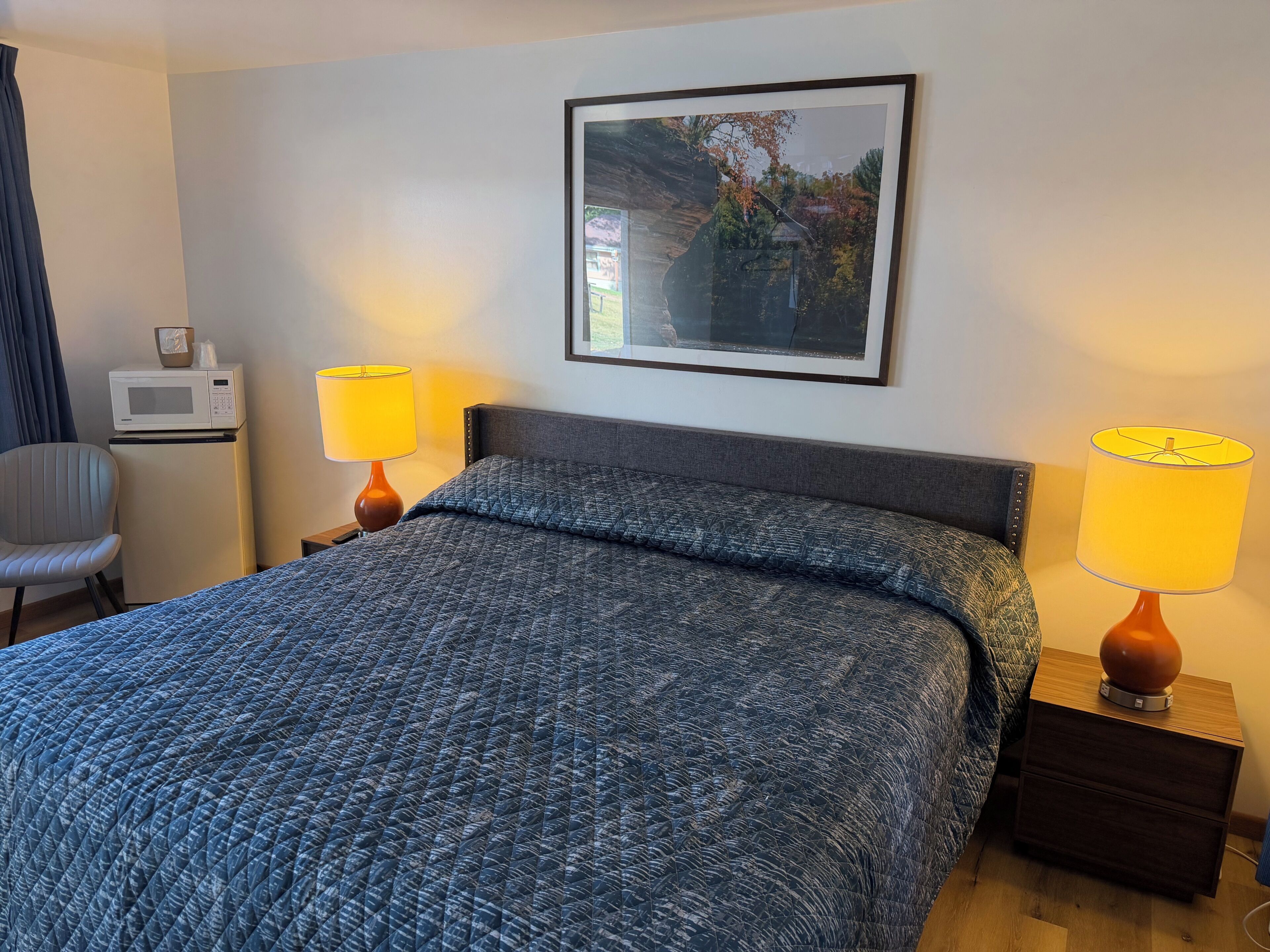 A hotel room with a bed, two bedside lamps, a microwave, a chair, and a framed picture on the wall.