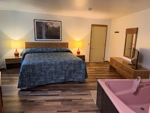 Quarto com cama, duas luminárias de cabeceira, cômoda de madeira, espelho grande, porta e banheira rosa.