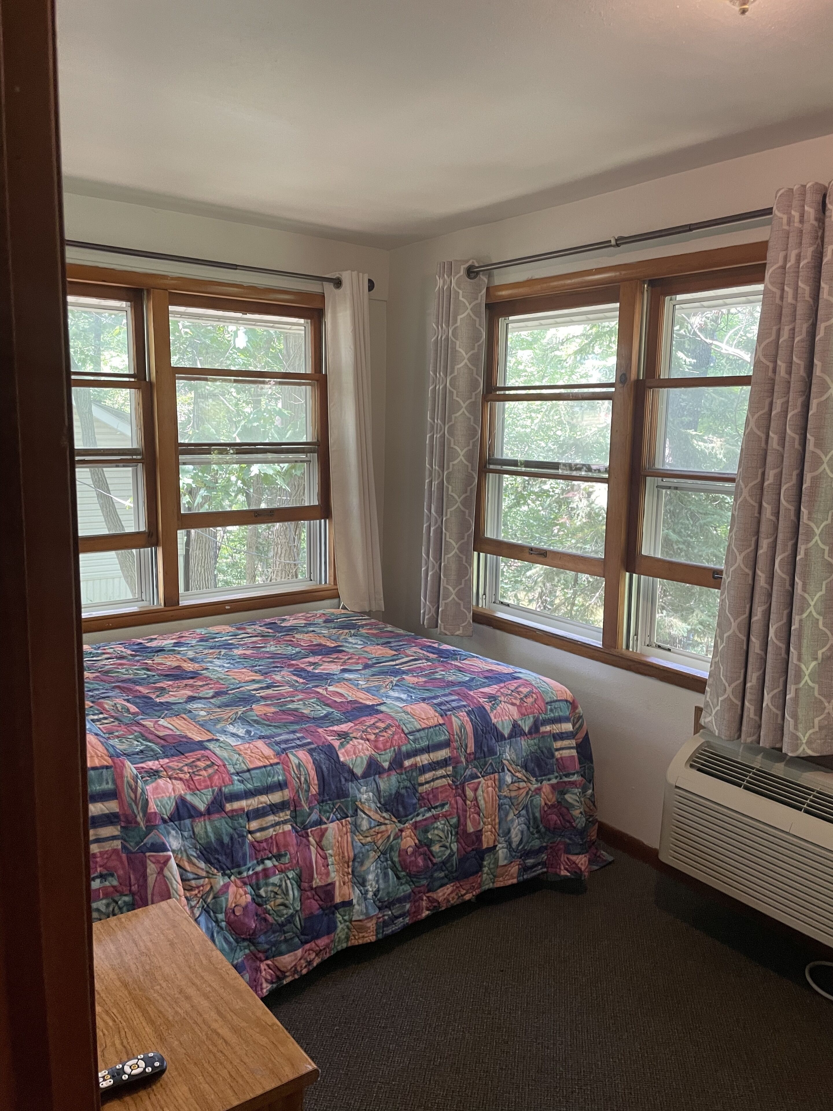 A bedroom with a large bed, two windows, a heater, and a wooden nightstand.