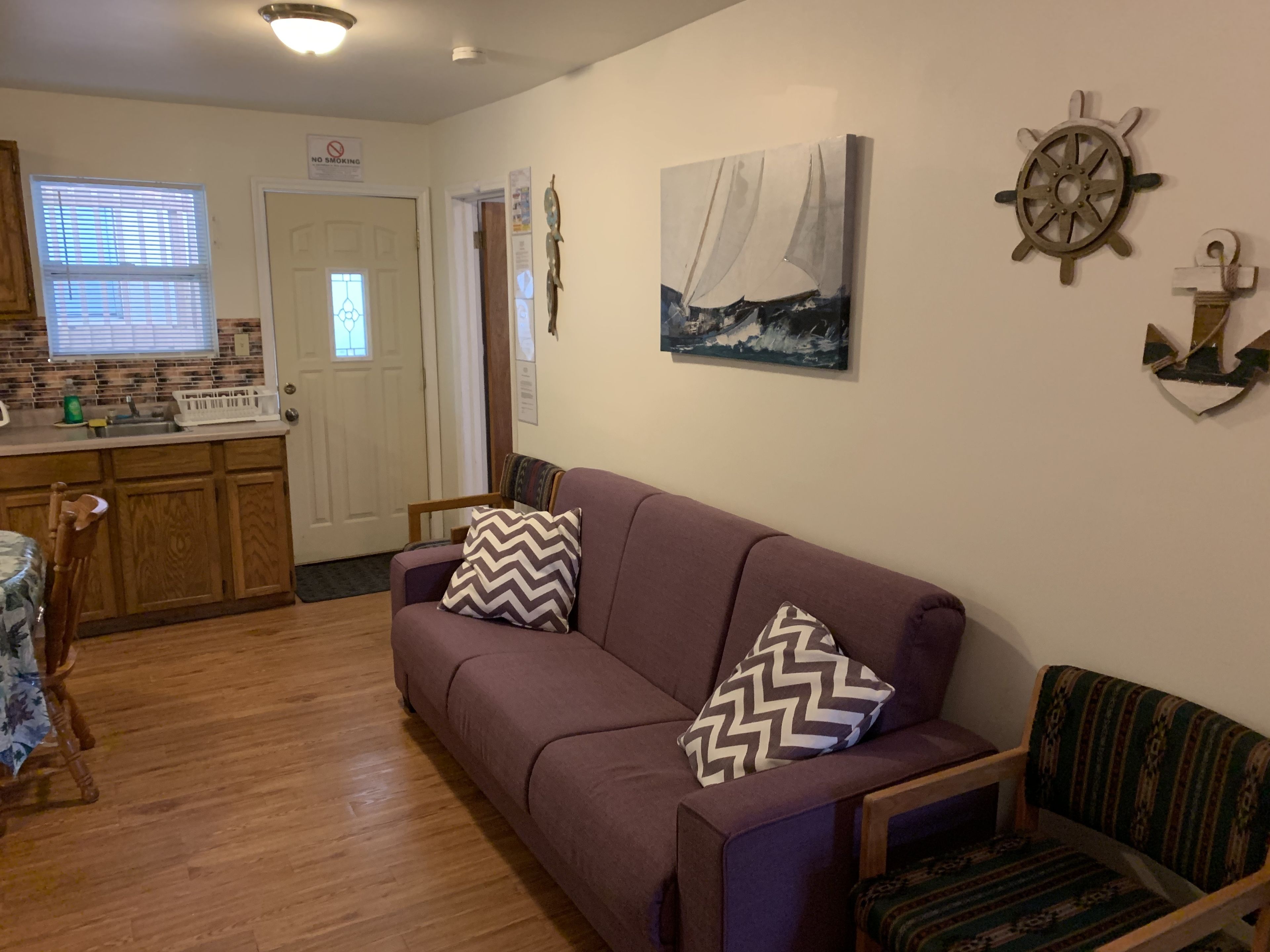 A living room with a purple sofa, wooden chairs, a wooden table, a kitchen area with a window, and nautical-themed wall decor.
