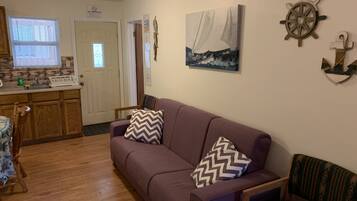 A living room with a purple sofa, wooden chairs, a wooden table, a kitchen area with a window, and nautical-themed wall decor.