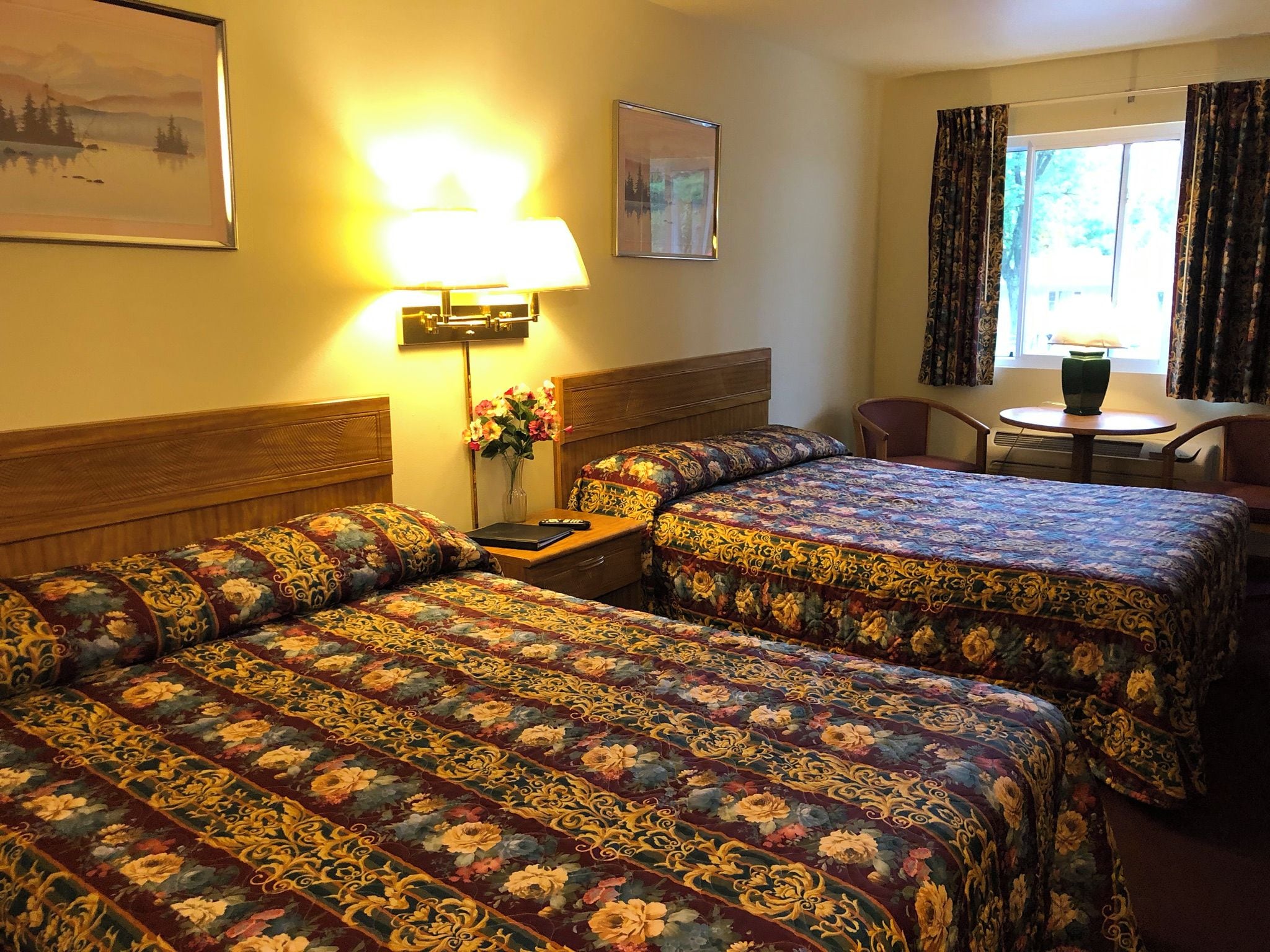 Habitación de hotel con dos camas, un escritorio, una silla, una lámpara y un ventanal con cortinas.