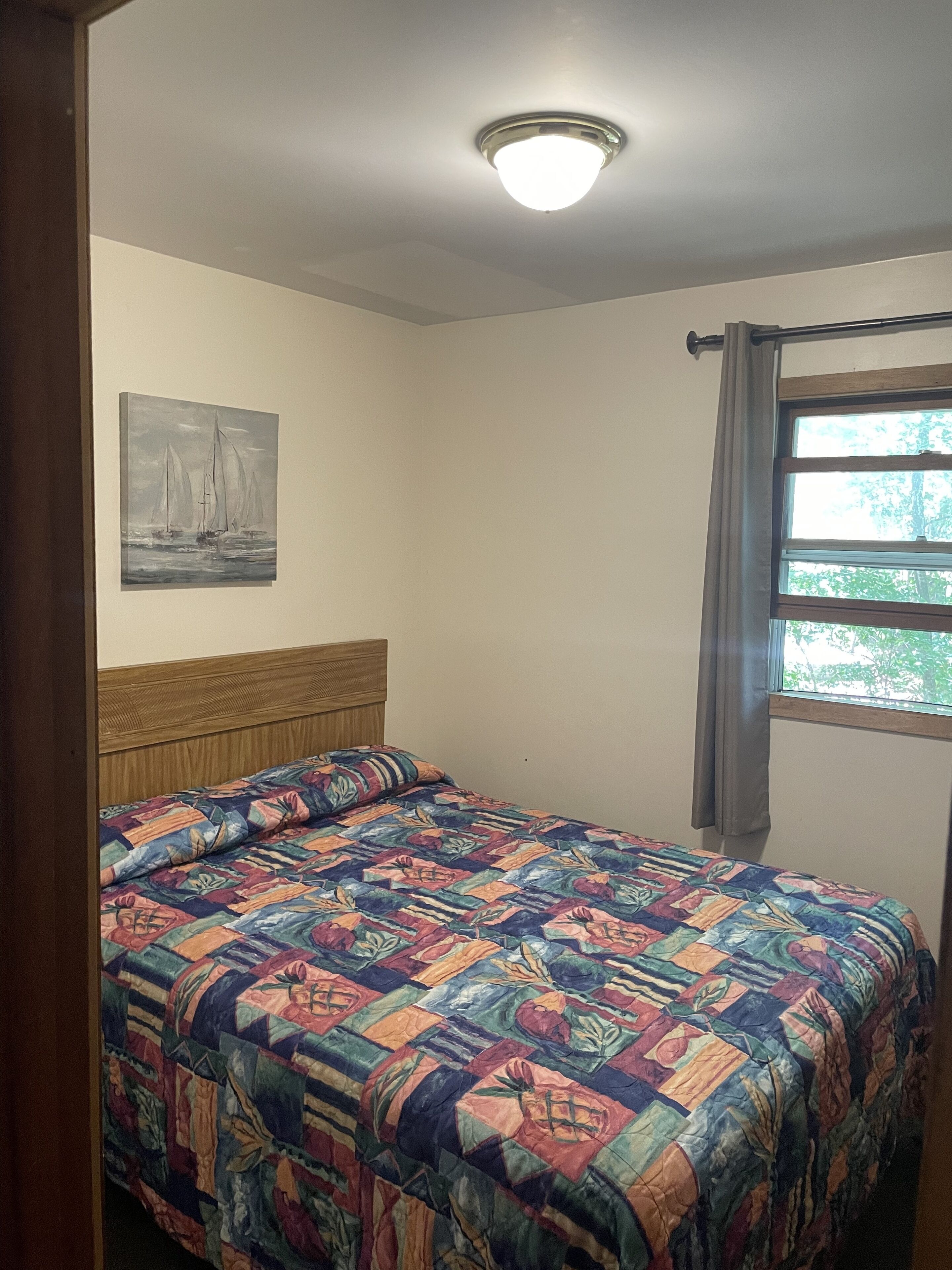 A bedroom with a wooden headboard, a colorful quilt, a painting of sailboats, and a window with curtains.