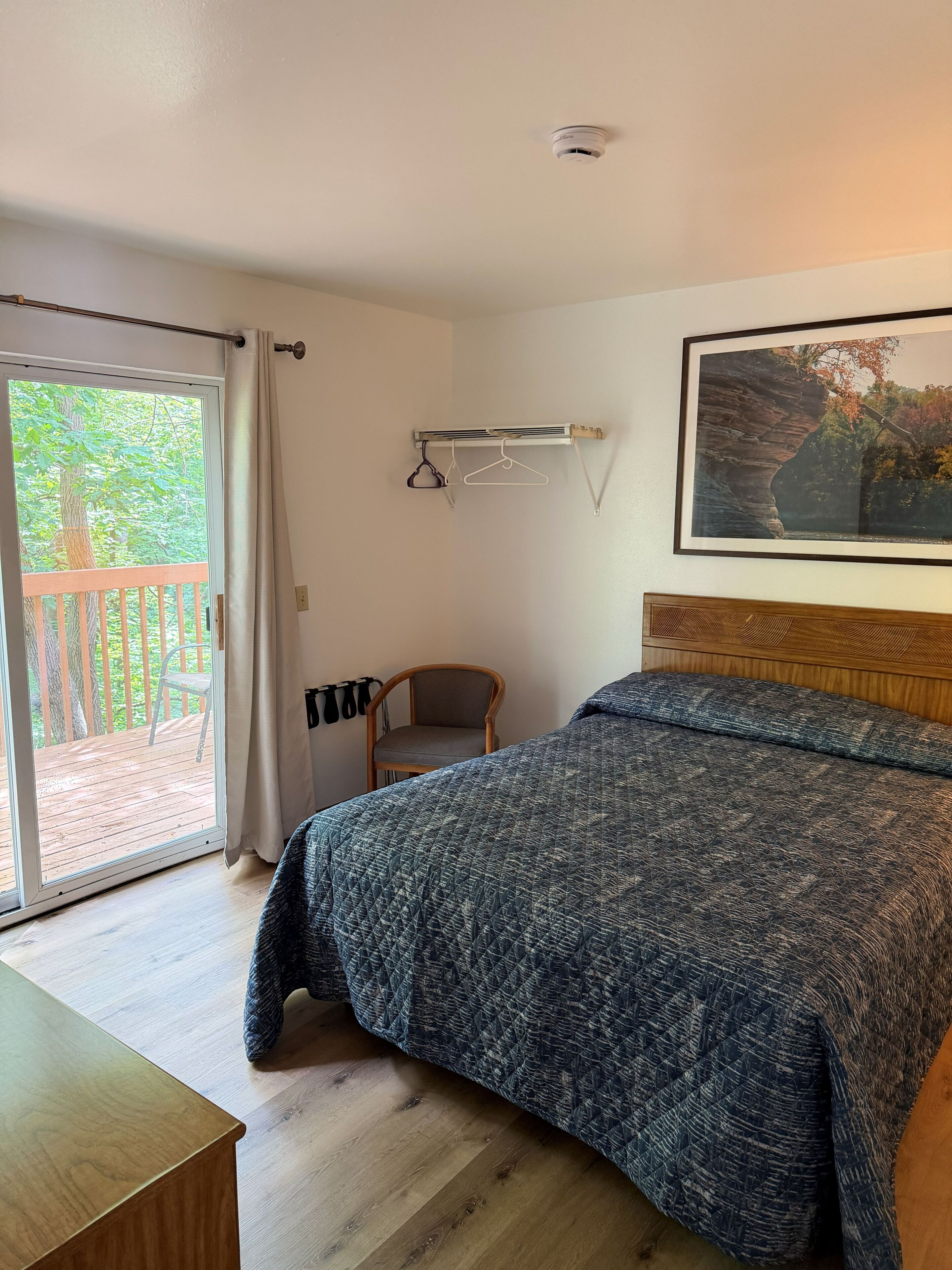 Ein Schlafzimmer mit einem Bett, einem Sessel, einem Kleiderhaken, einem Bild an der Wand und einer Schiebetür zum Balkon.