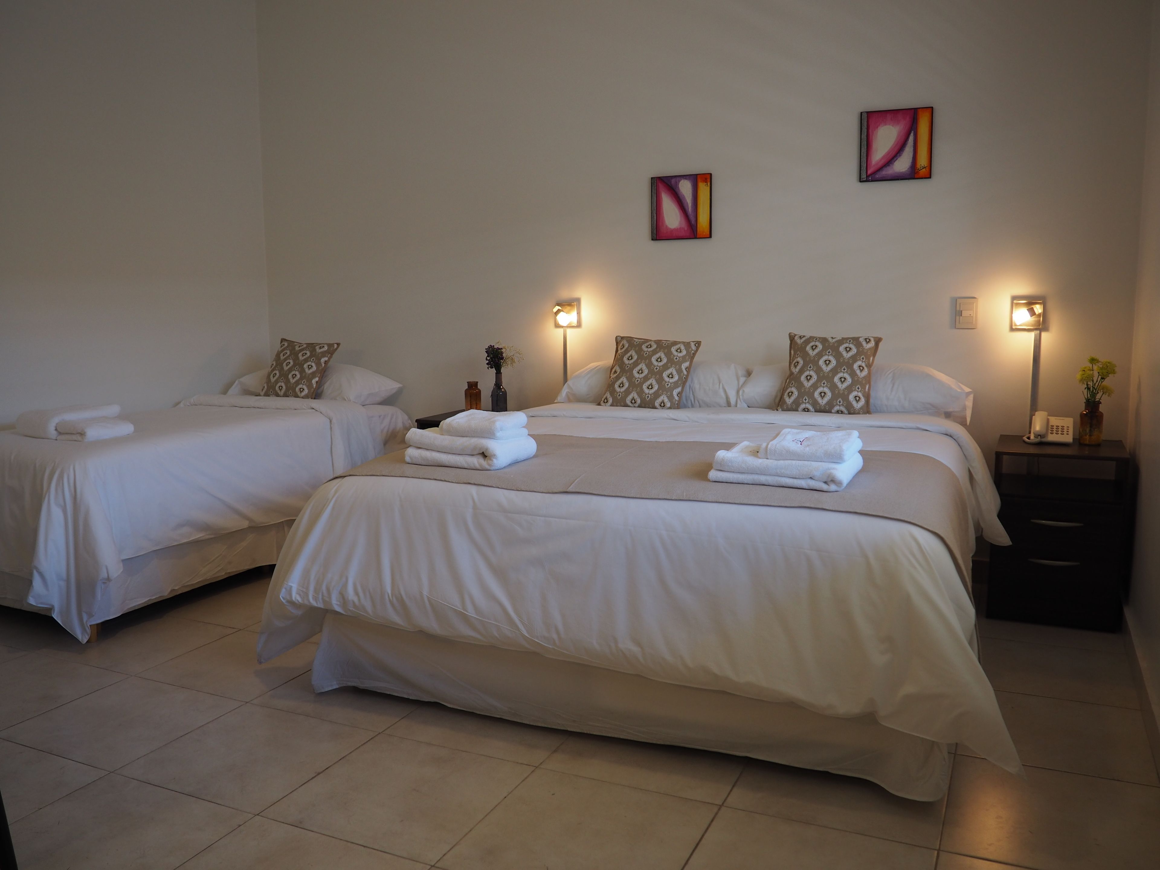 Habitación de hotel con dos camas, cada una con sábanas y almohadas blancas, una mesita de noche con lámpara, y una obra de arte en la pared.