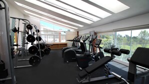 Un gimnasio con diversos equipos de ejercicio, ventanas amplias y vistas al exterior con vegetación.