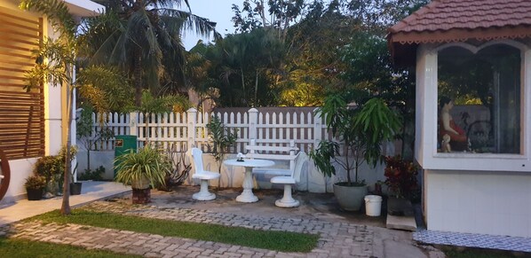 Un patio con mobiliario blanco de exterior, una cerca blanca de piquetes y varias macetas con plantas.