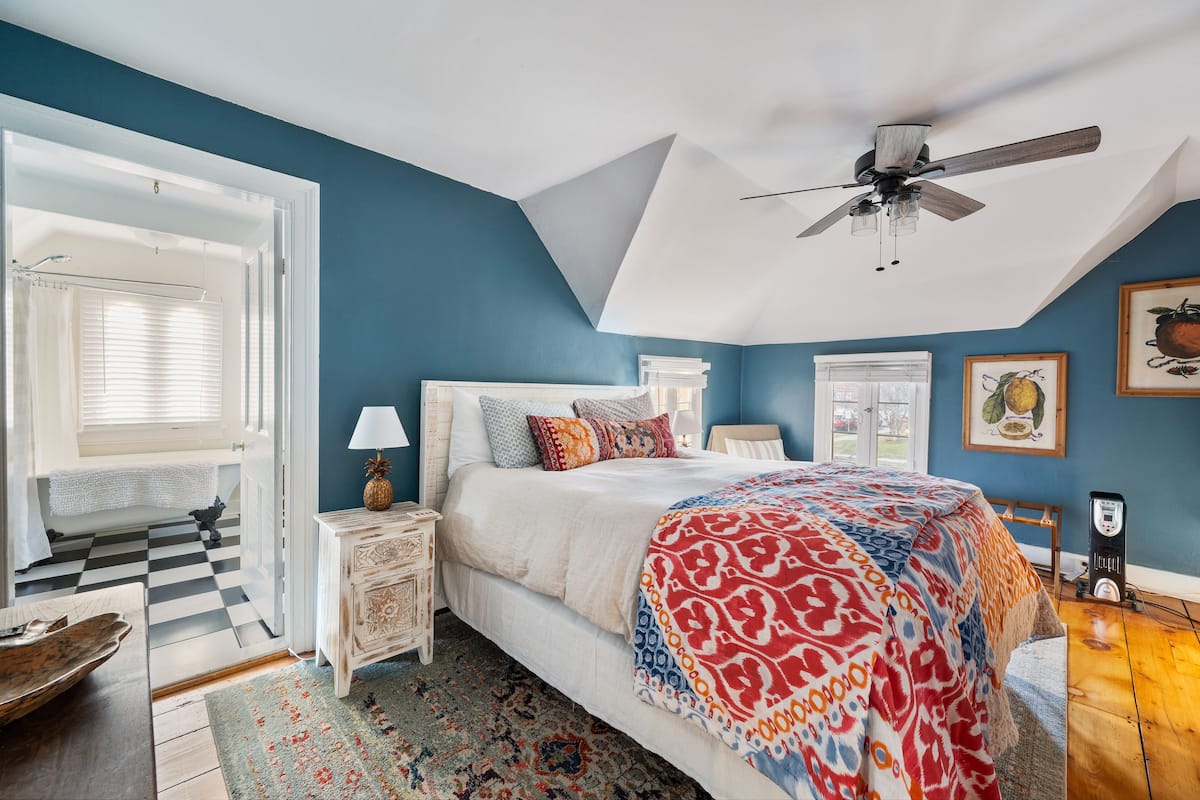 A bedroom with a bed, a ceiling fan, a nightstand, a lamp, a heater, and a window with blinds.