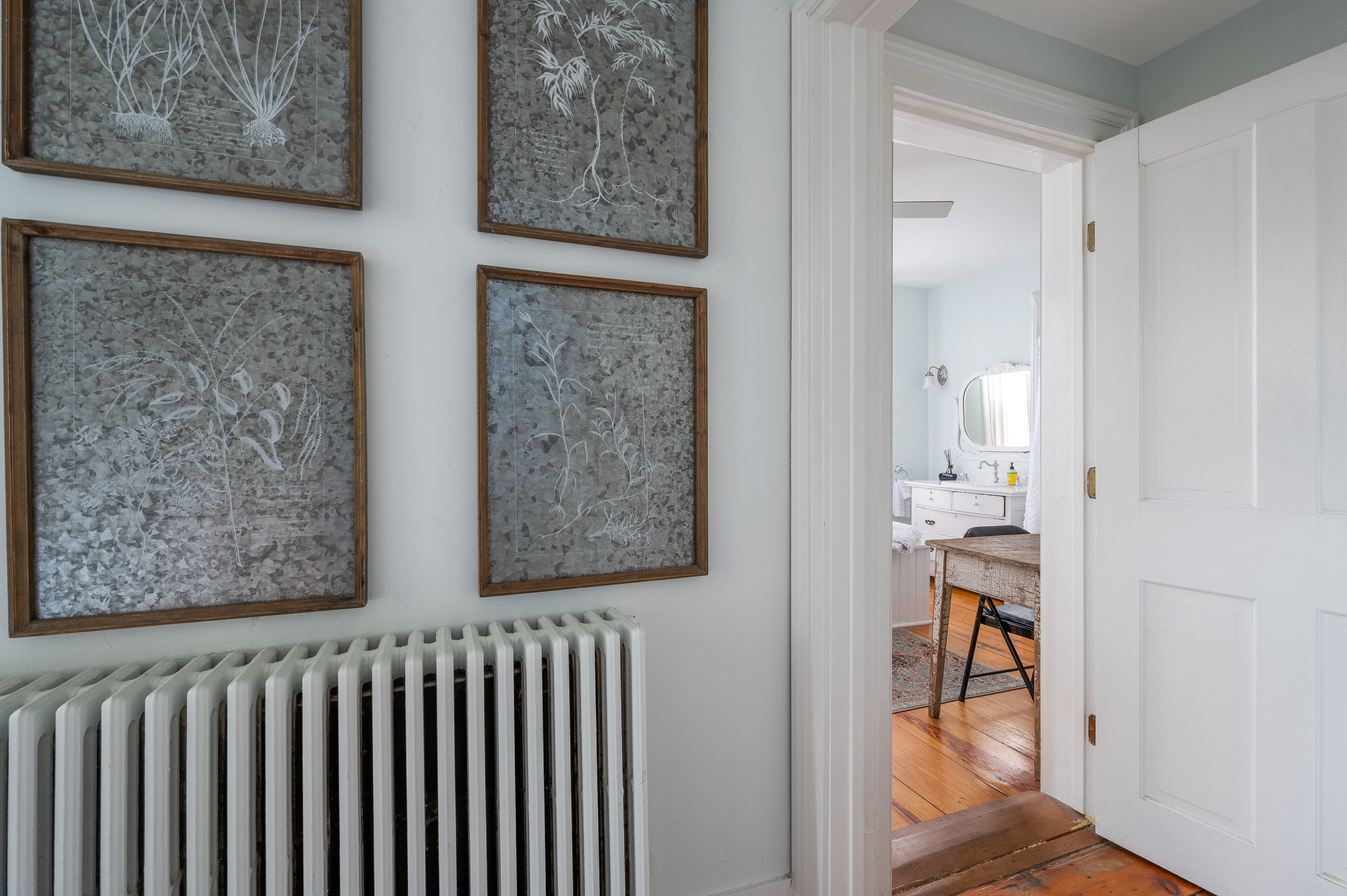 A hallway with four framed abstract wall art pieces, a white radiator, and a wooden floor leading to a room with a white door.
