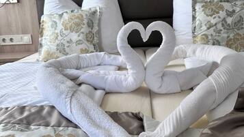 A neatly made bed with a heart-shaped towel arrangement, floral-patterned pillows, and a wooden headboard.