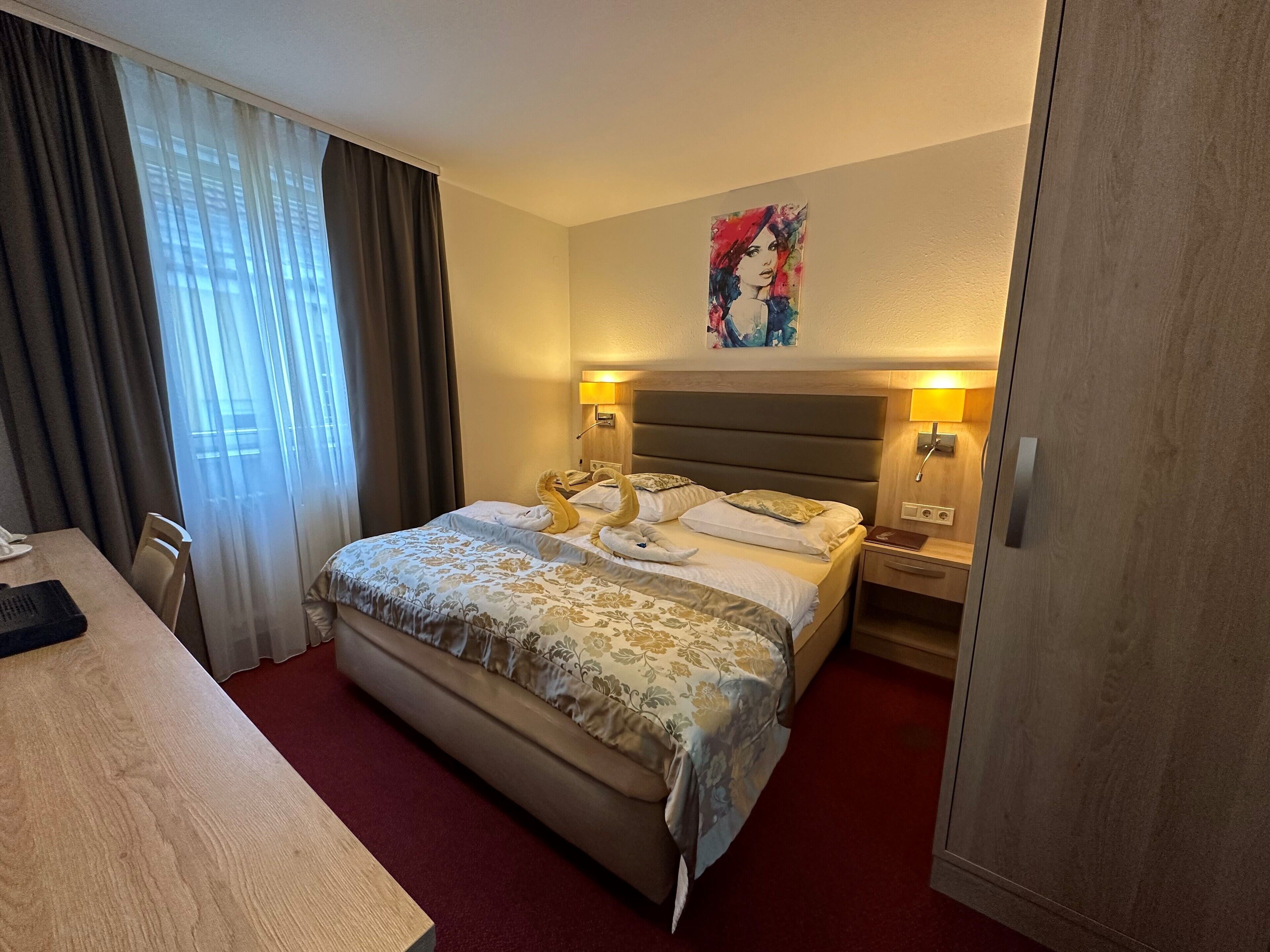 A hotel room with a bed, a desk, a chair, and a painting on the wall.